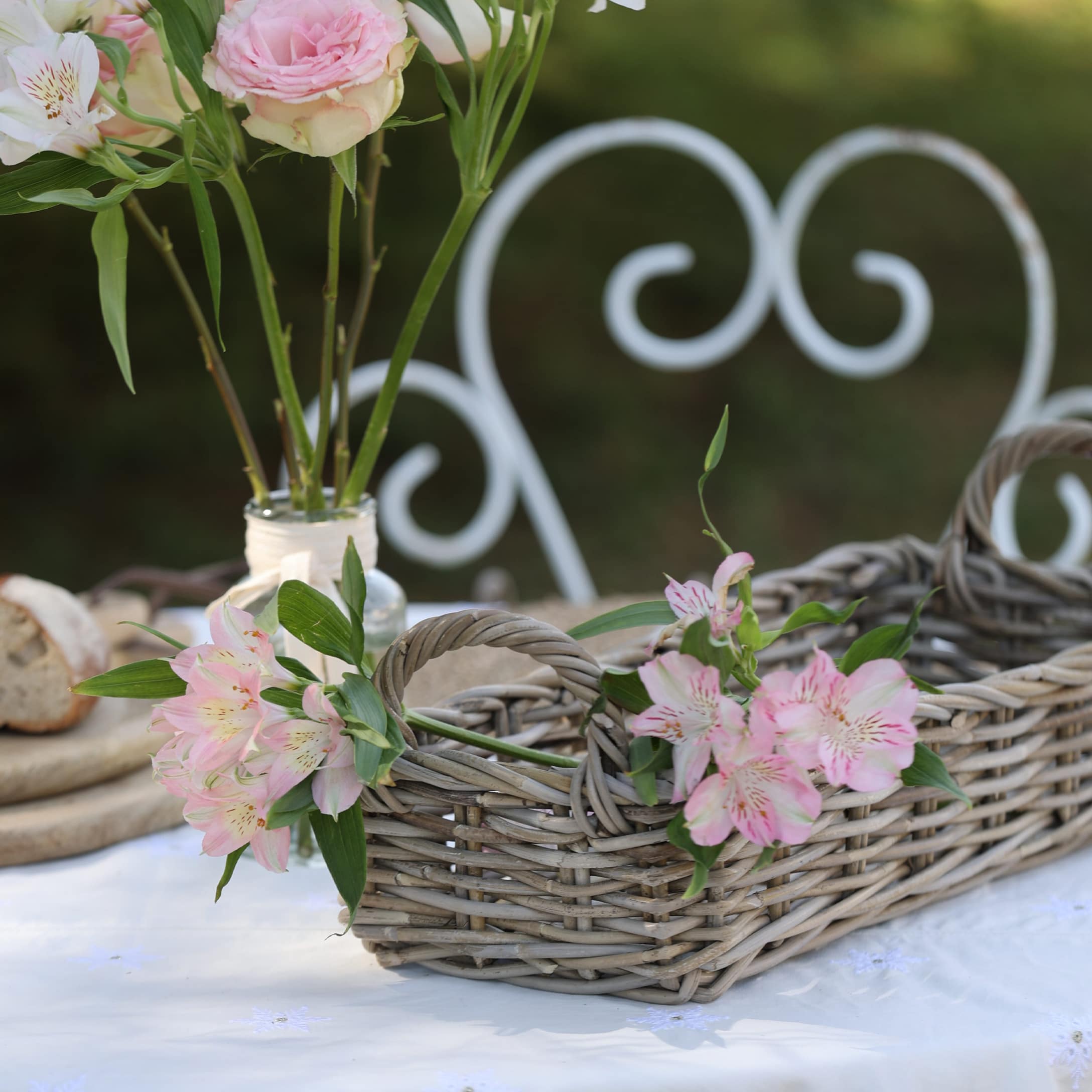 Rustikaler grauer Weidenkorb mit rosa Blumen auf weisser Tischdecke im Garten