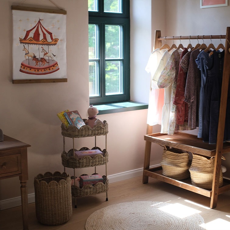 Dreistöckiges Korbregal mit Spielzeug und Kinderkleidung an Holzkleiderständer in hellem Zimmer