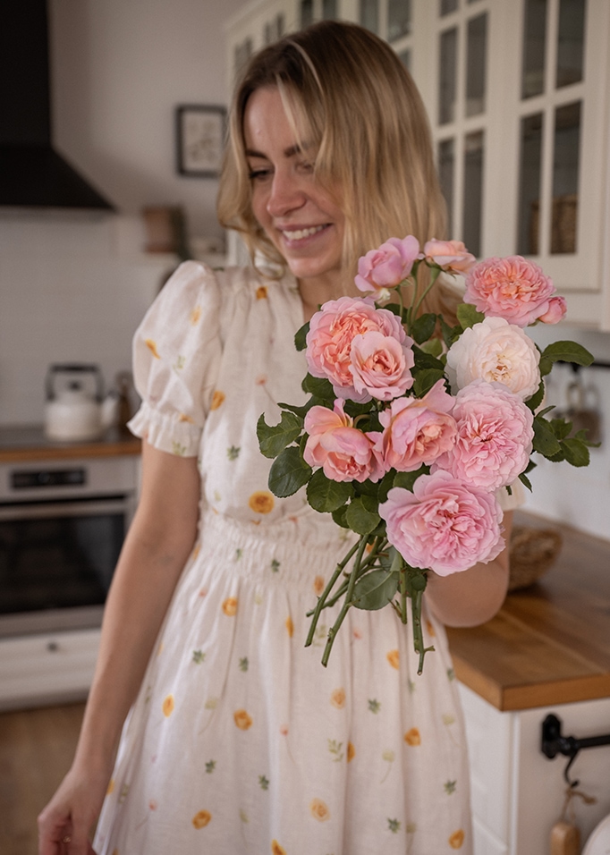 Frau im weissen Blumenkleid hält einen Strauß rosa Rosen in moderner Küche.