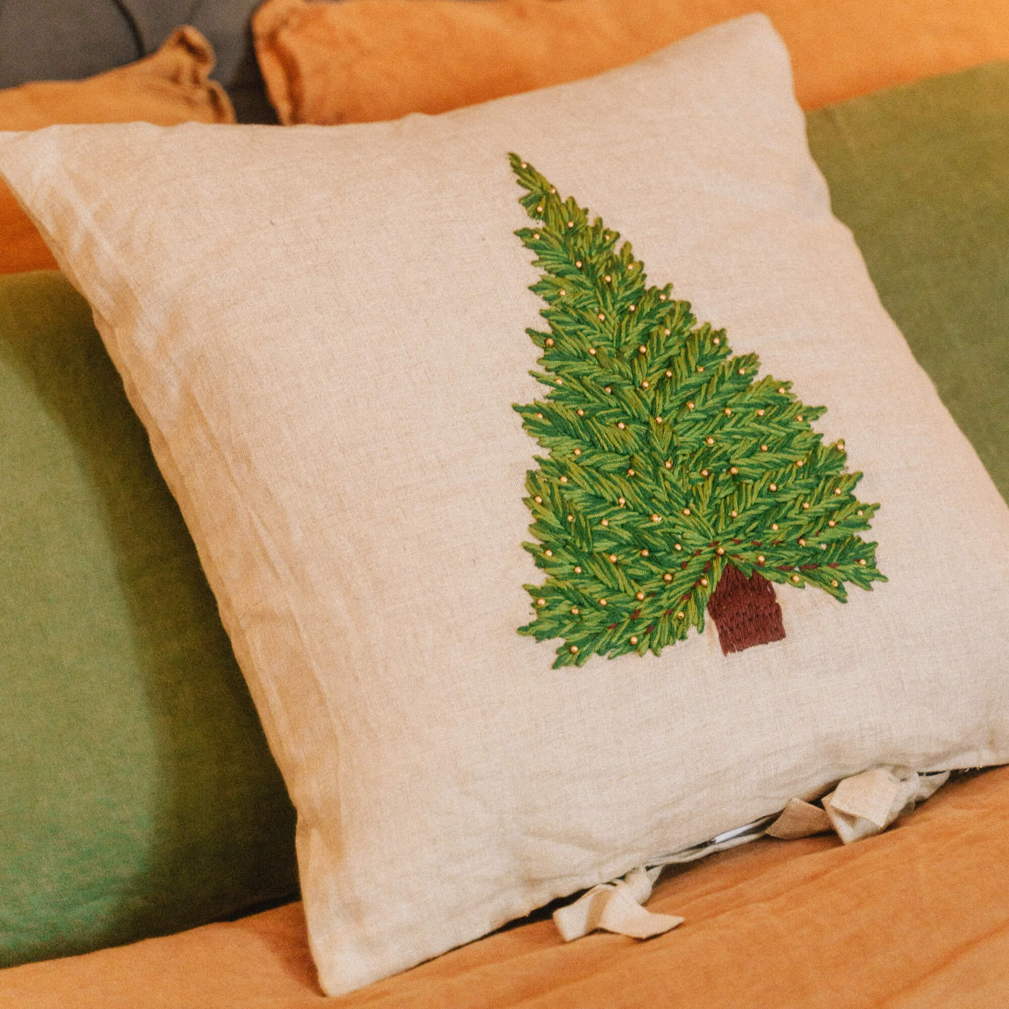 Beiges Leinenkissen mit grün besticktem Tannenbaum auf orange-grüner Bettwäsche
