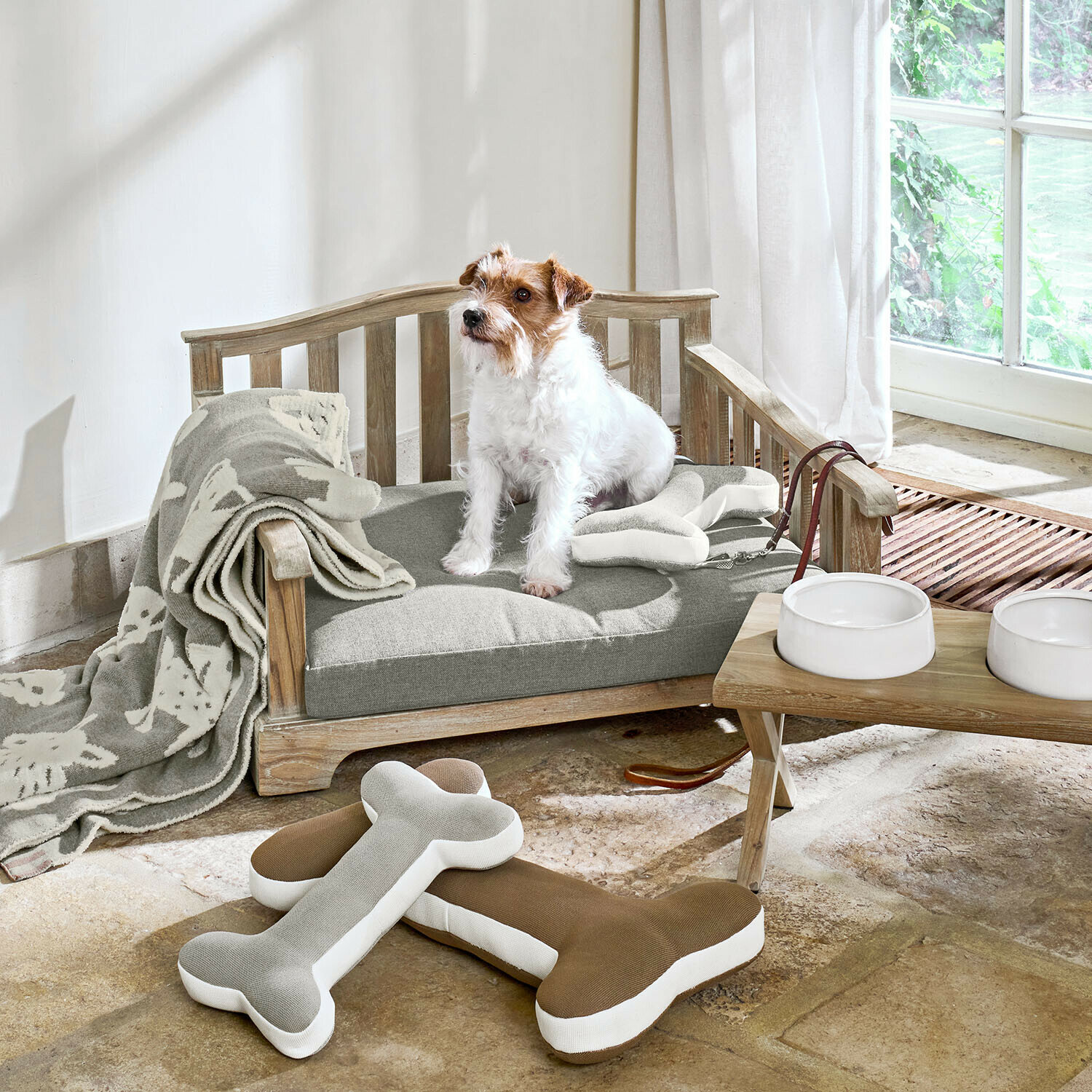 In- & Outdoor-Haustiersofa Waggins Holz-Hundebett mit grauem Kissen, Knochen-Spielzeug, helle Decke und weissen Futternäpfen am Fenster