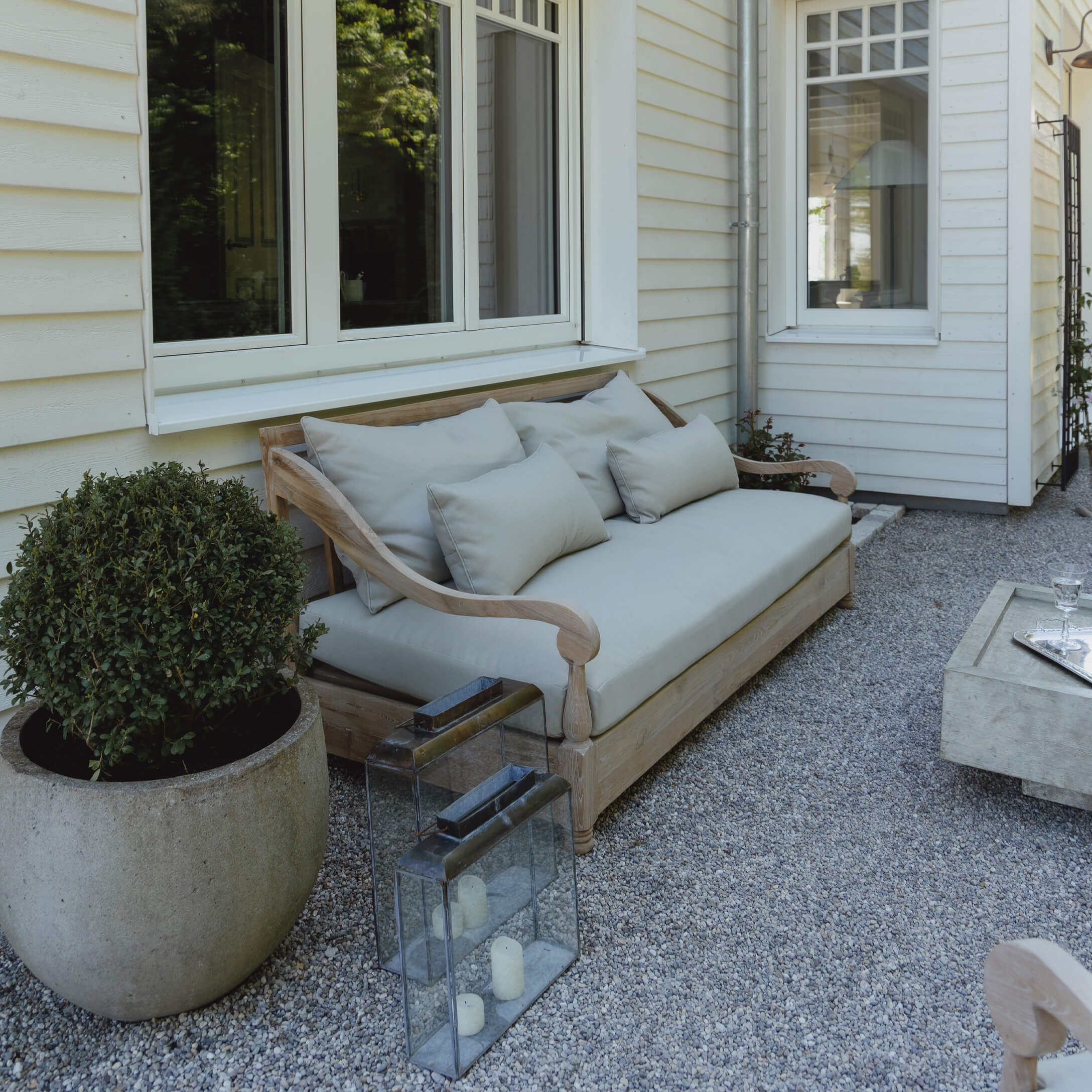 Holz-Gartensofa mit hellgrauen Kissen auf Kiespatio vor weissem Haus