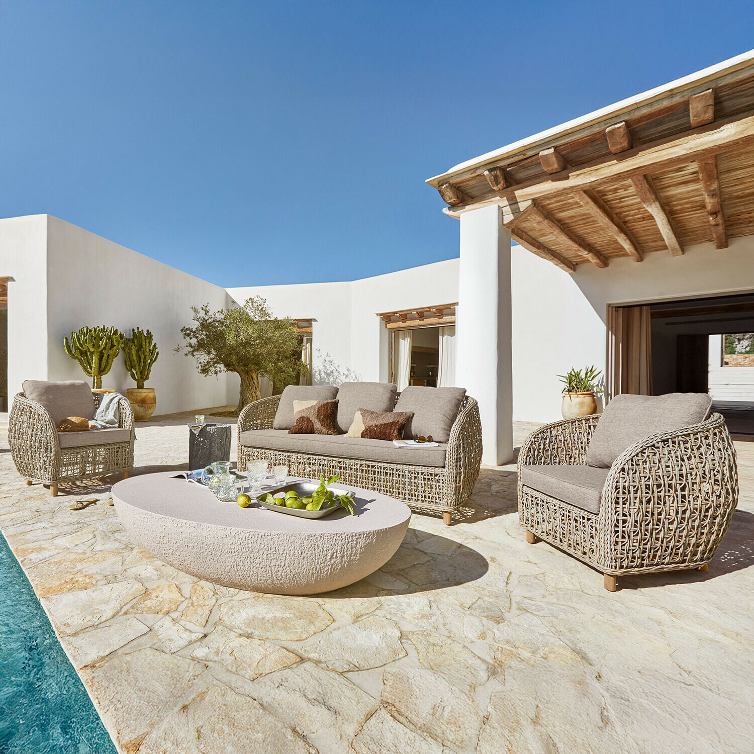 Beiges Rattan-Gartenmöbel-Set mit Polstern auf Stein-Terrasse neben Pool bei Sonnenschein