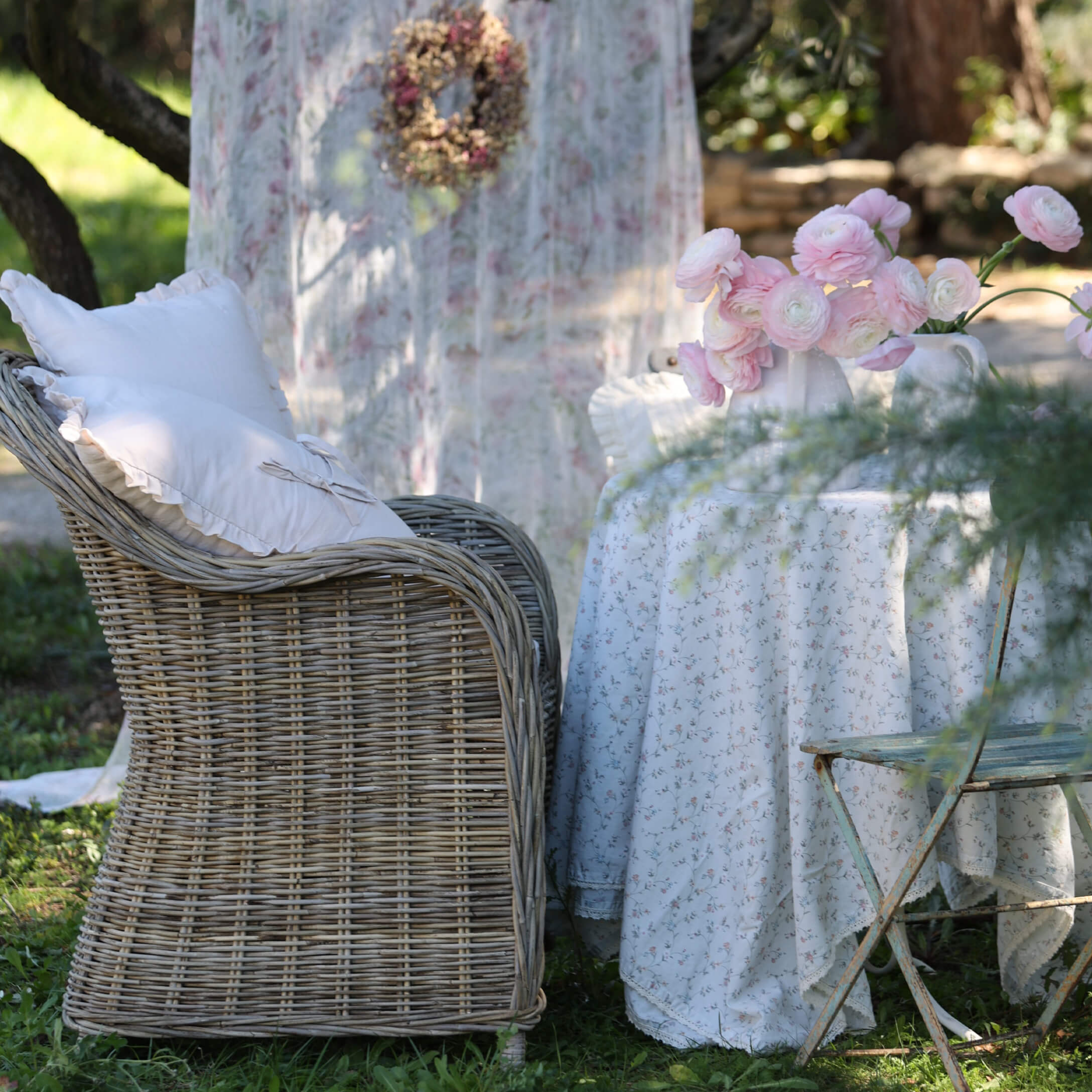 Wiklinersessel mit weissen Kissen neben Tisch mit floralem Tischtuch und rosa Blumen im Garten