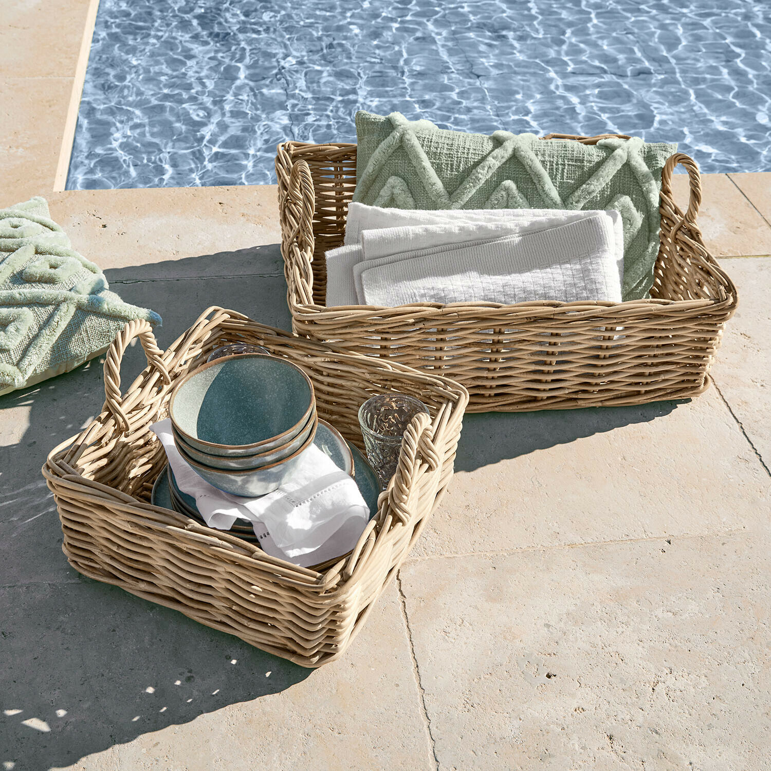 Zwei geflochtene Körbe mit Griffen, gefüllt mit grünen Kissen und weissen Handtüchern am Poolrand
