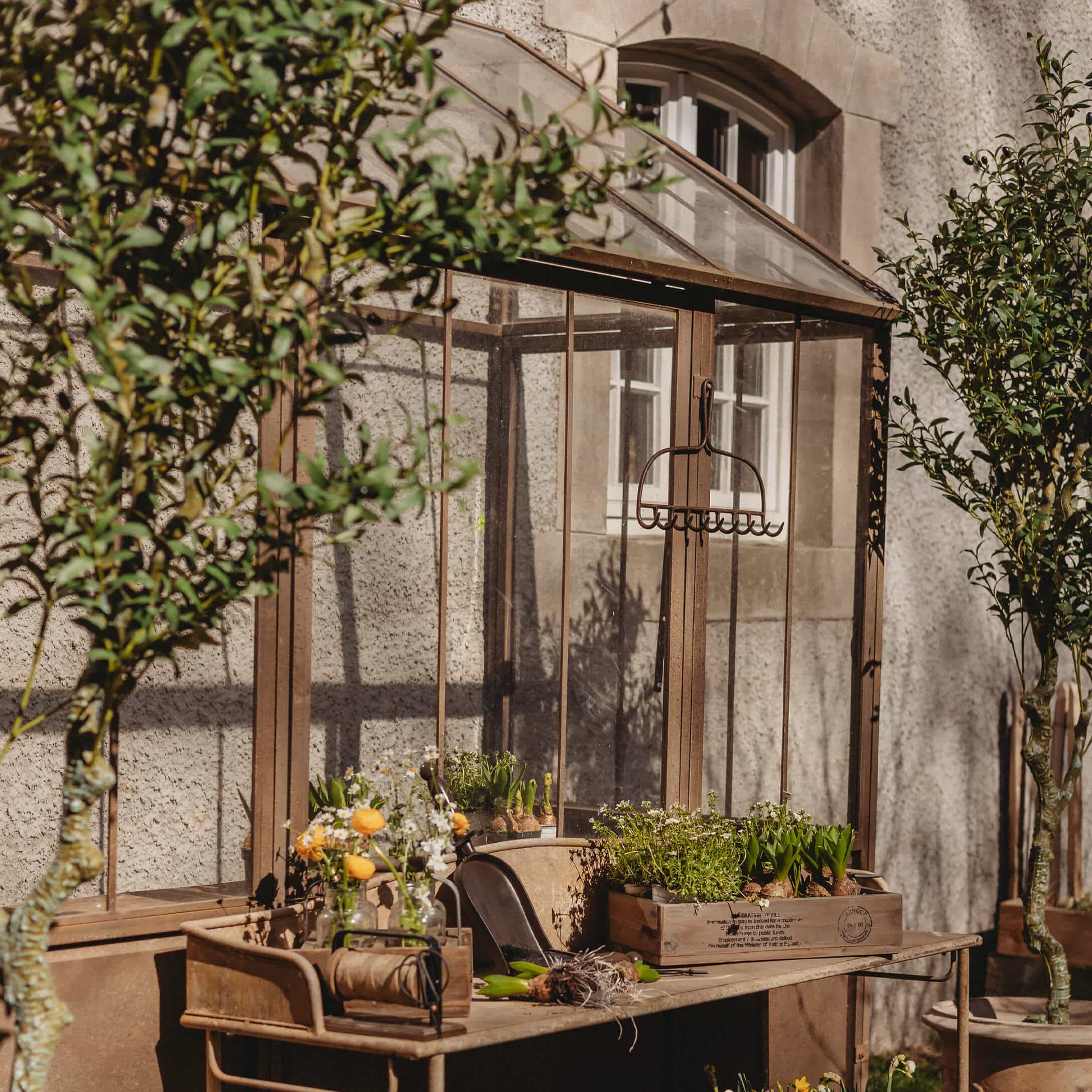 Vintage Metall-Gartentisch mit Blumentöpfen und Werkzeugen vor einem Gewächshaus