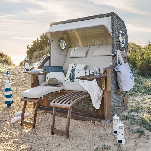 Strandkorb aus Rattan mit beigen Polstern und Holzfussstützen im Sand, ideal zum Entspannen