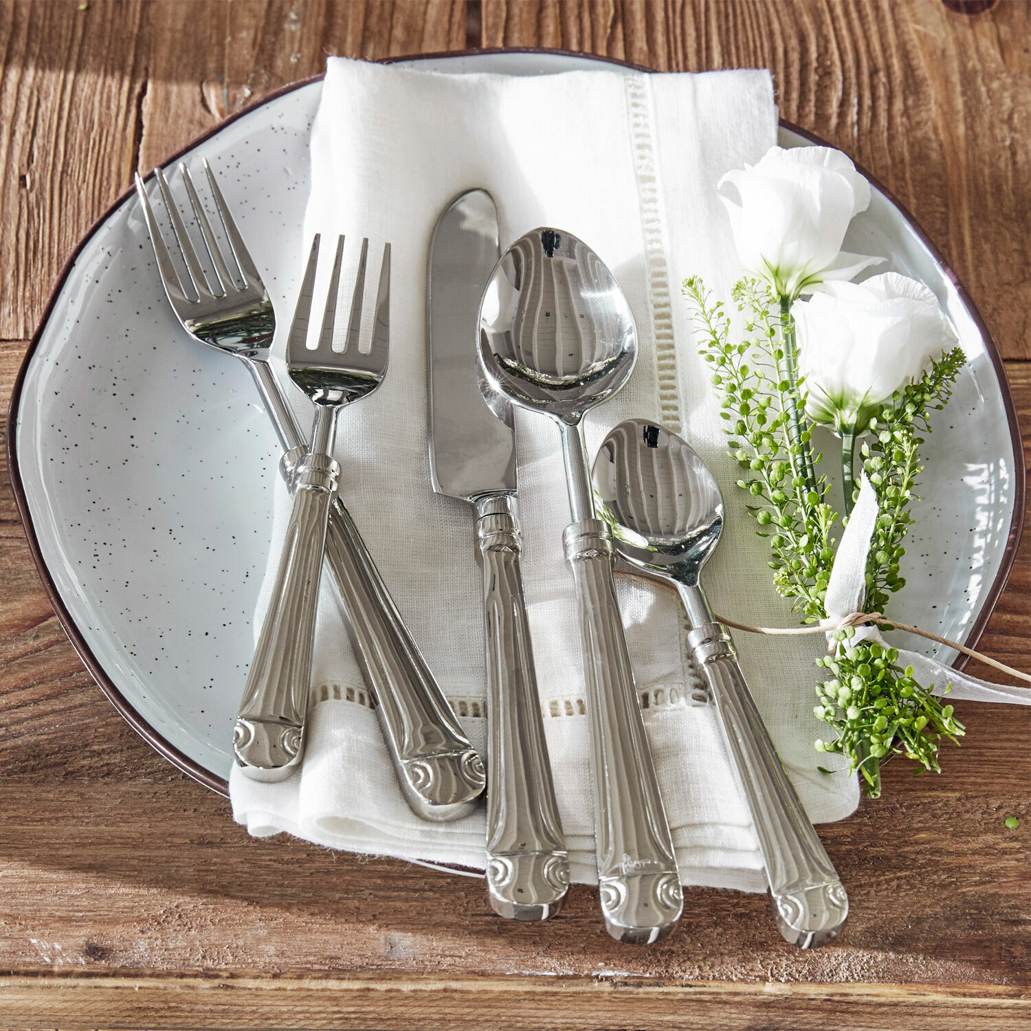 Besteck Eriat Set aus silbernem Besteck auf weissem Stoff mit Blumenarrangement auf Holztisch