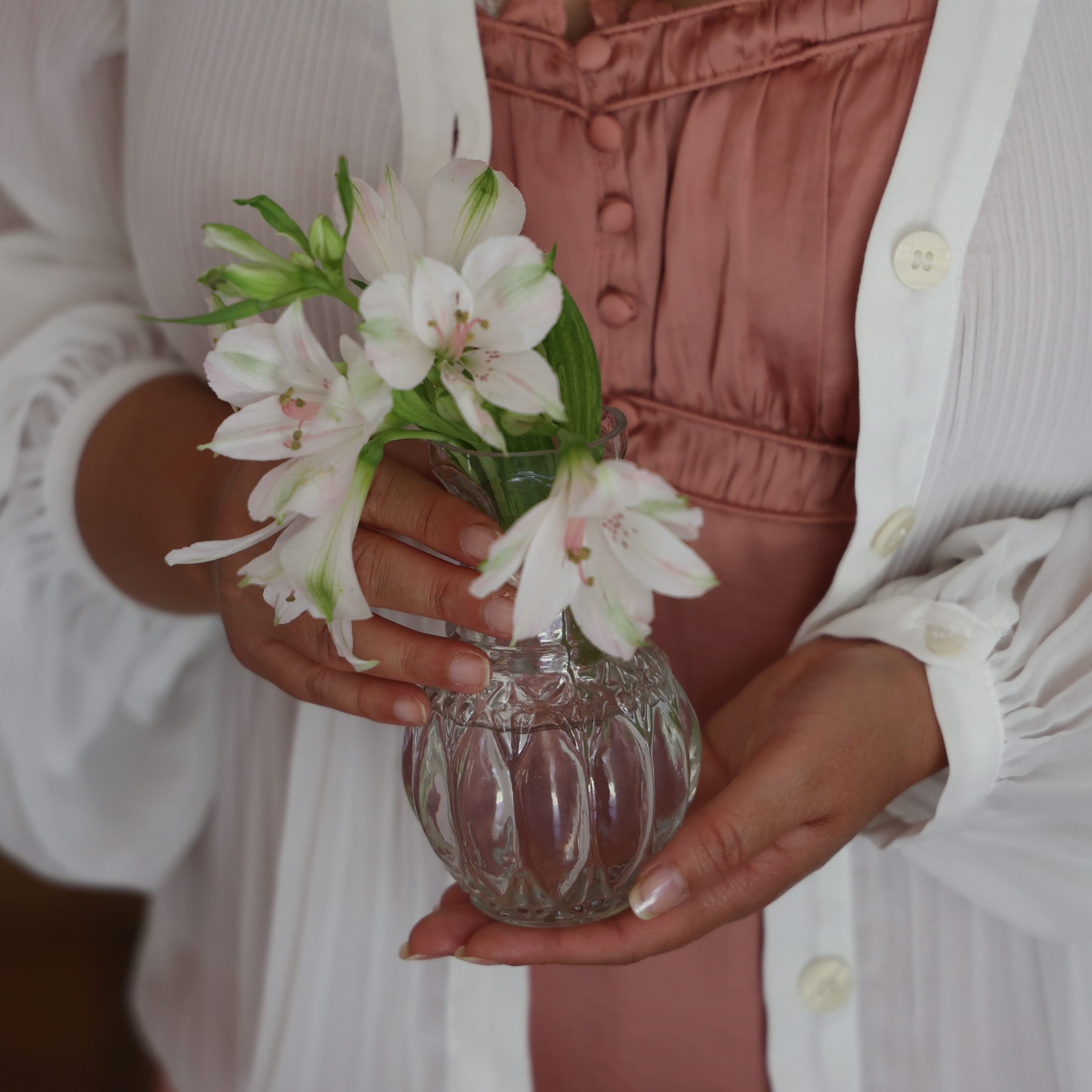 Glasvase mit weissen Blumen, gehalten von einer Person in weissem Cardigan und rosa Kleid