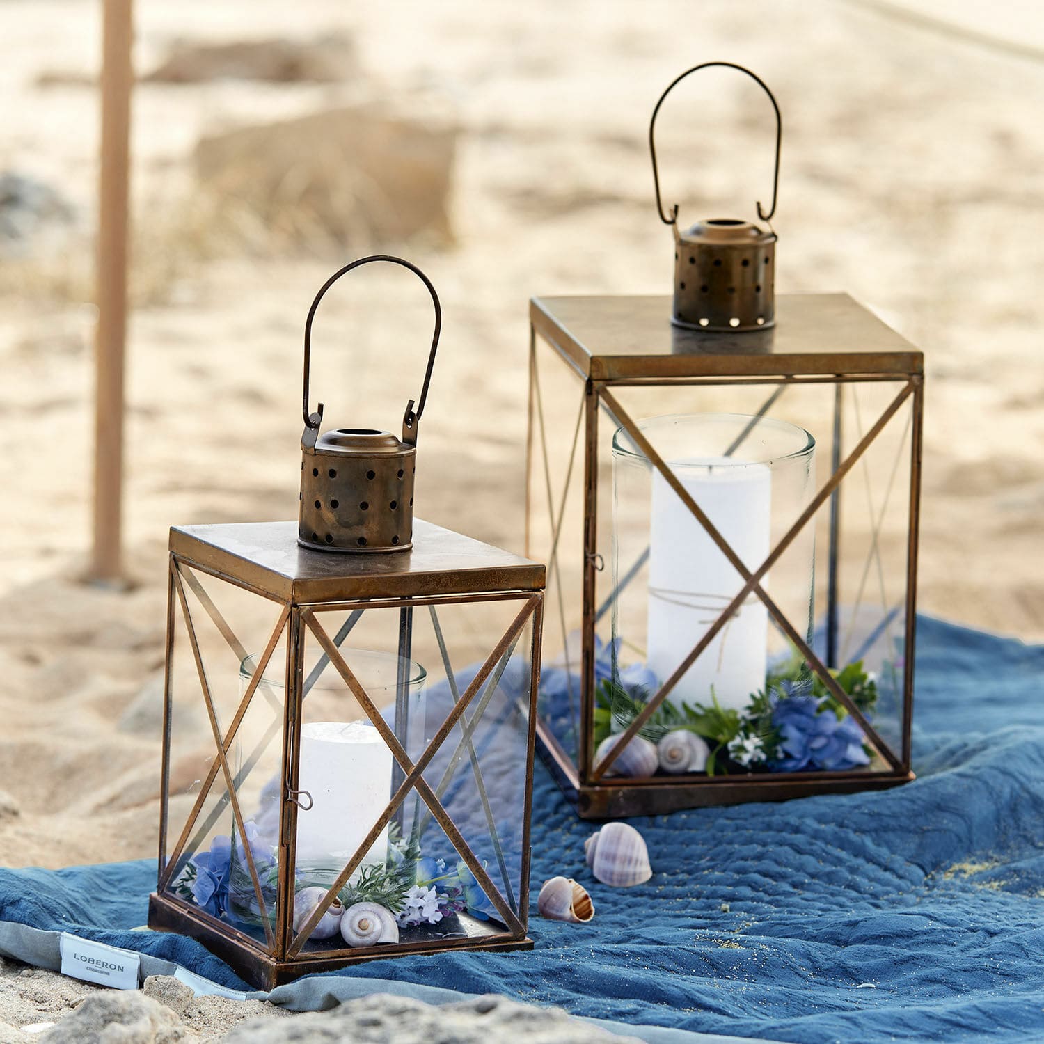 Laterne 2er Set Garance Zwei braune Metalllaternen mit weissen Kerzen und Dekorationen auf blauem Stoff am Strand