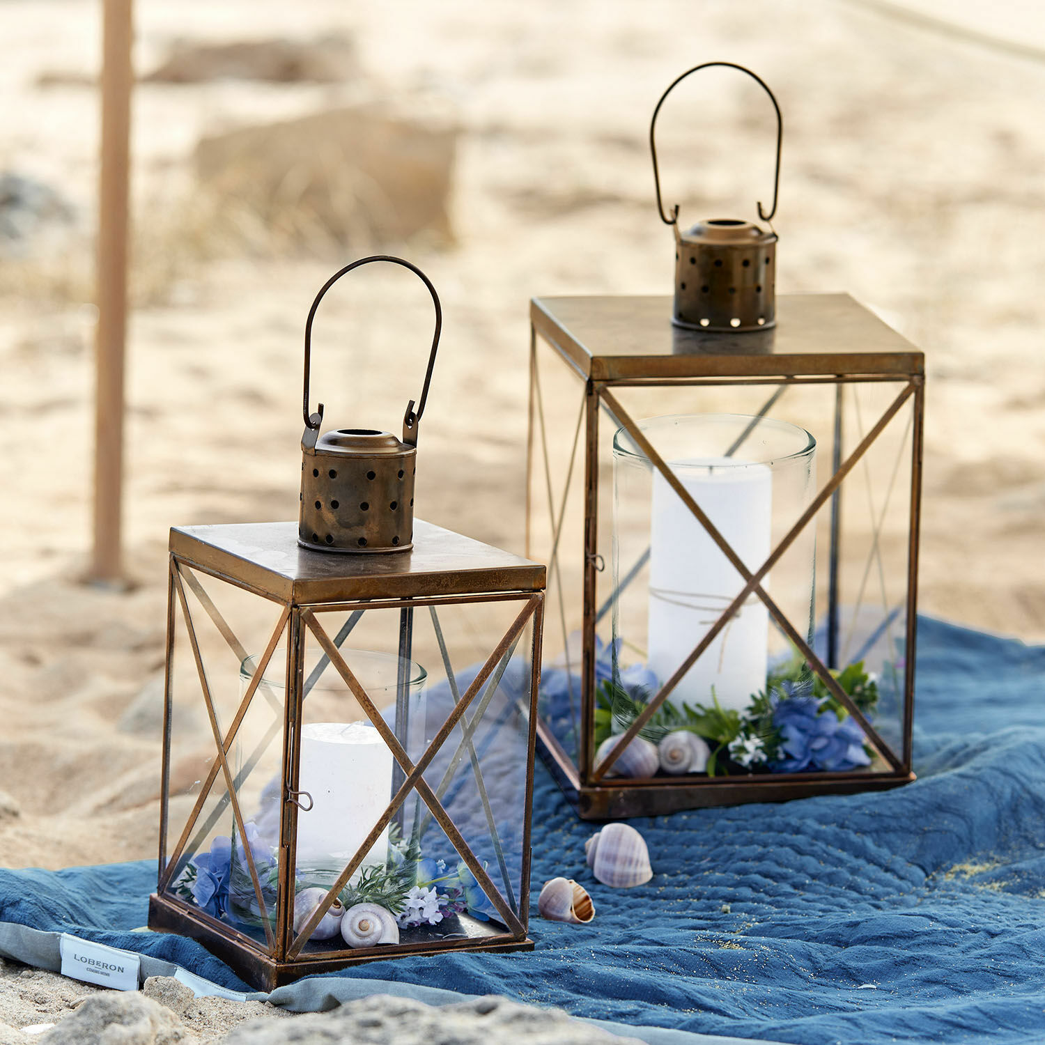 Laterne 2er Set Garance Zwei braune Metalllaternen mit weissen Kerzen und Dekorationen auf blauem Stoff am Strand