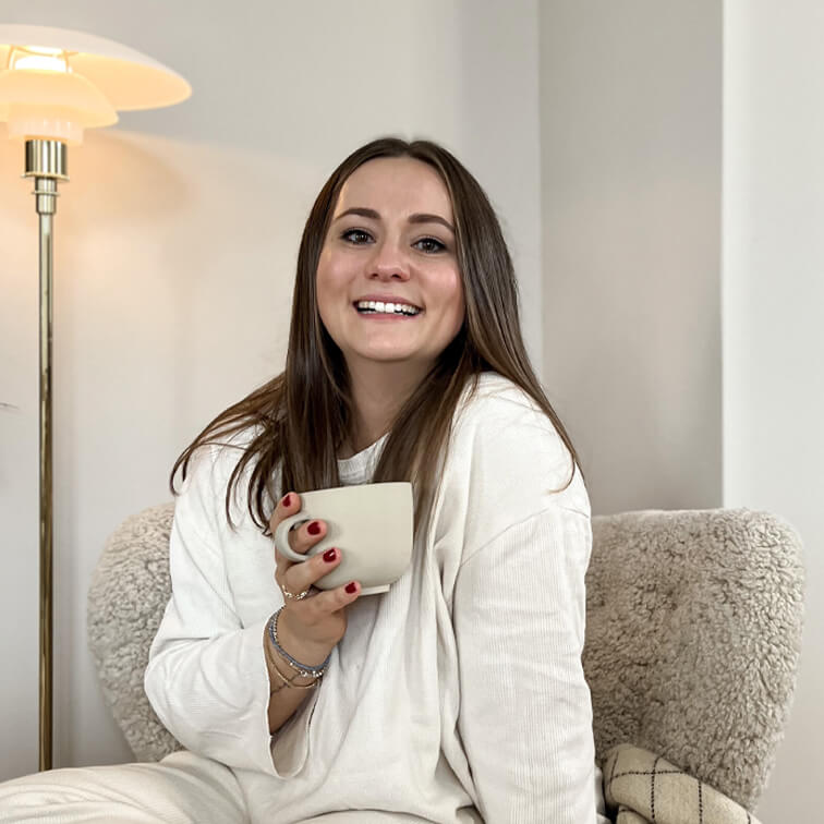 Lächelndi Frau im wiisse Pullover hält e beige Tasse und sitzt iinem gmüetliche Sessel im warme Raum