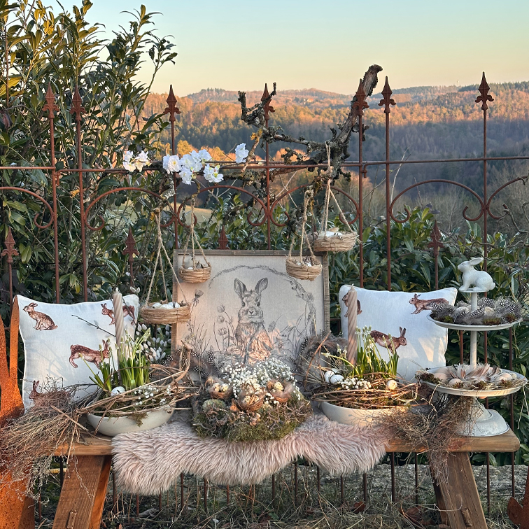 Rustikale Osterdekoration mit Hasenkissen, Weidenkörben und natürlichen Blumen auf einer Holzbank
