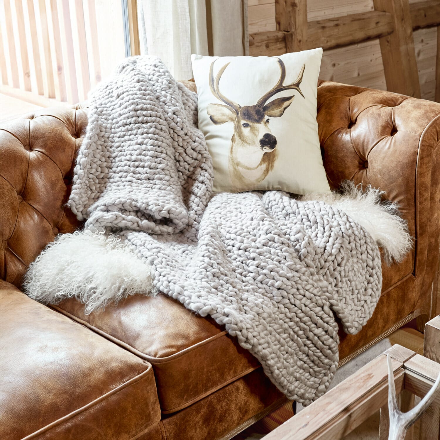 Plaid Reidous Braunes Ledersofa mit grob gestrickter grauer Decke und Kissen mit Hirschmotiv in rustikalem Zimmer