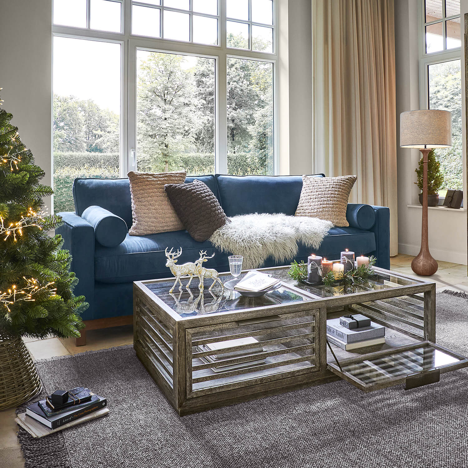 Gemütliches Wohnzimmer mit dunkelblauem Sofa, Glastisch und weihnachtlicher Dekoration mit Tannenbaum und Kerzen
