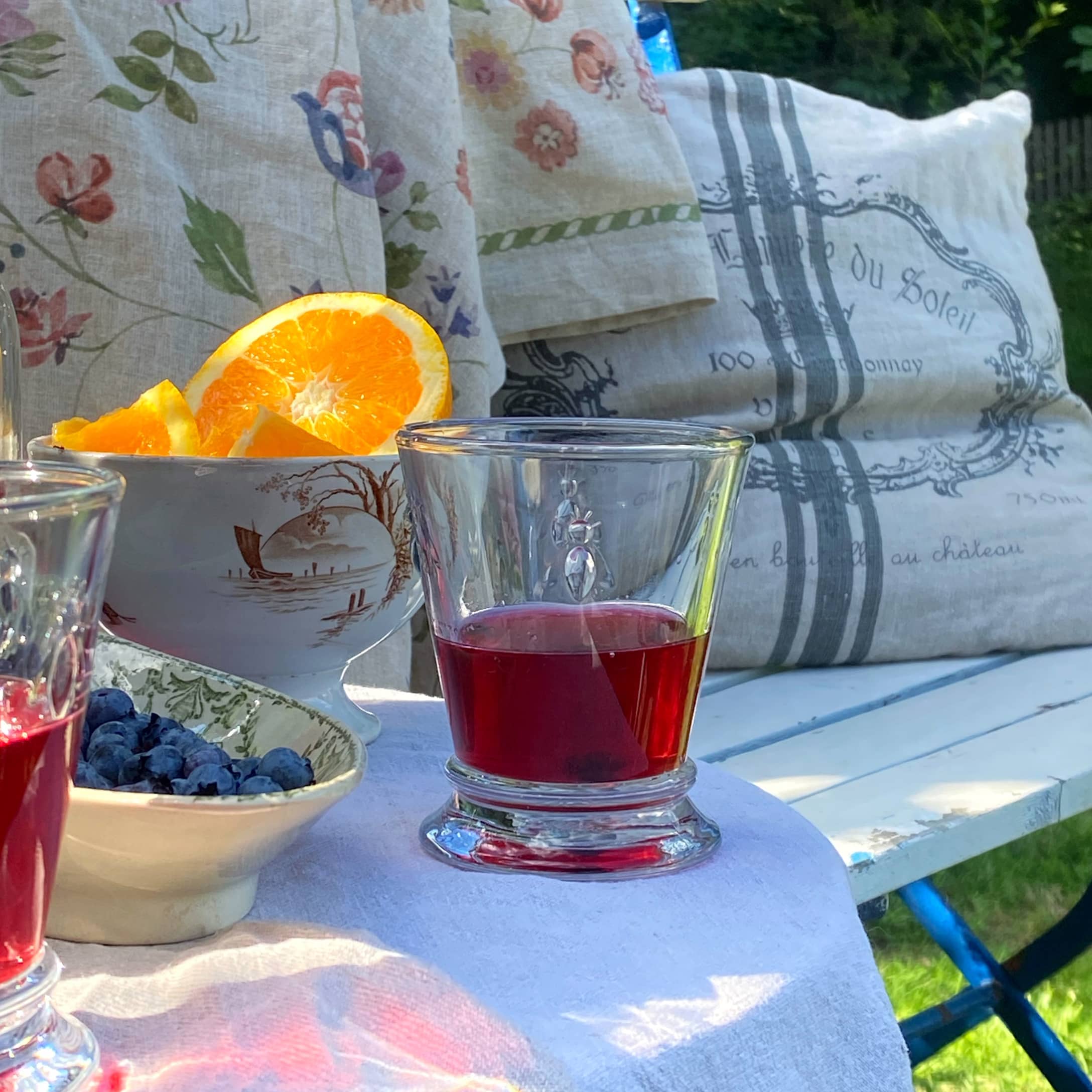 Glas mit rotem Getränk auf weissem Tuch mit Früchten und Kissen im Garten