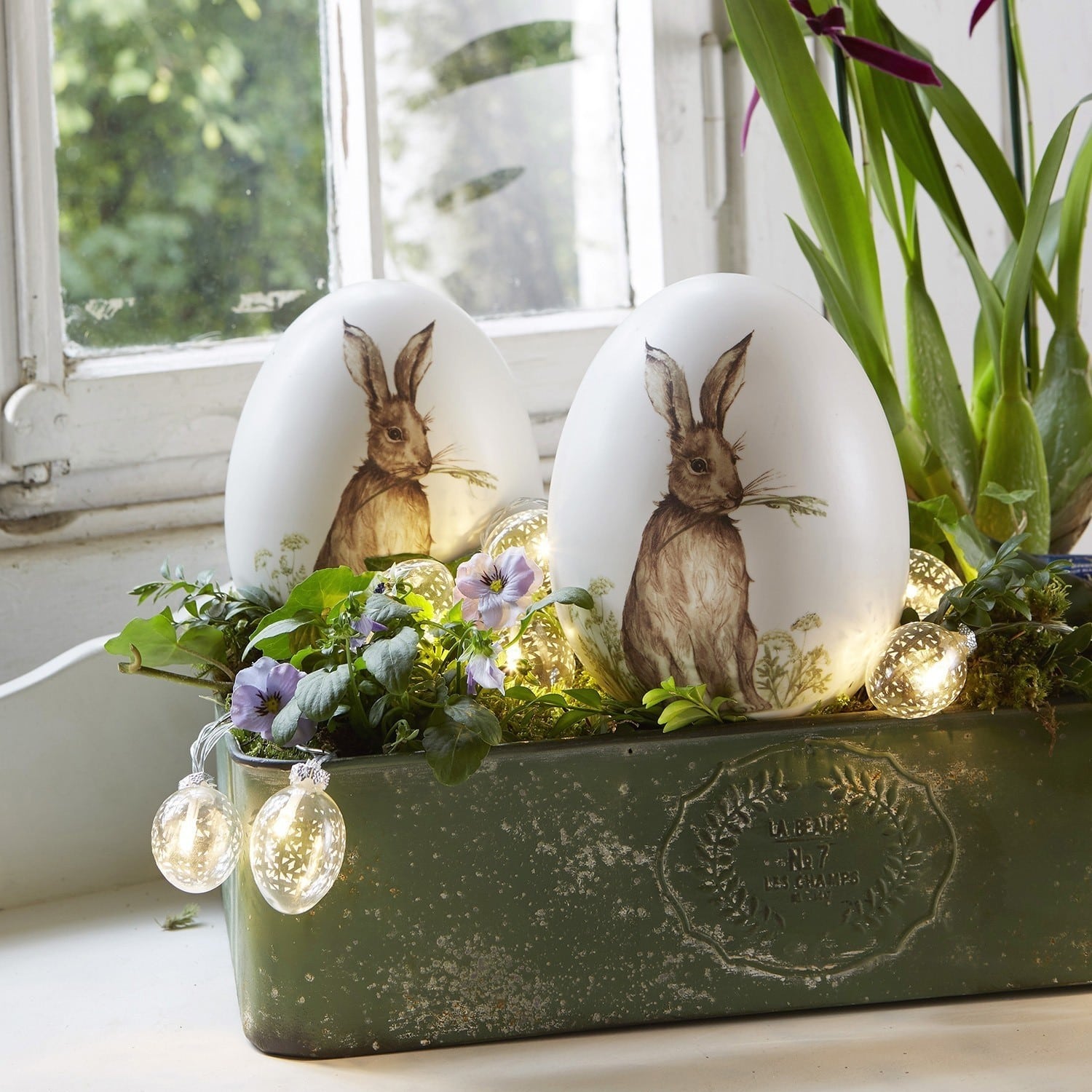 Weisse Dekoeier mit Hasenmotiv in grünem Pflanzgefäss mit Blumen und LED-Lichterkette