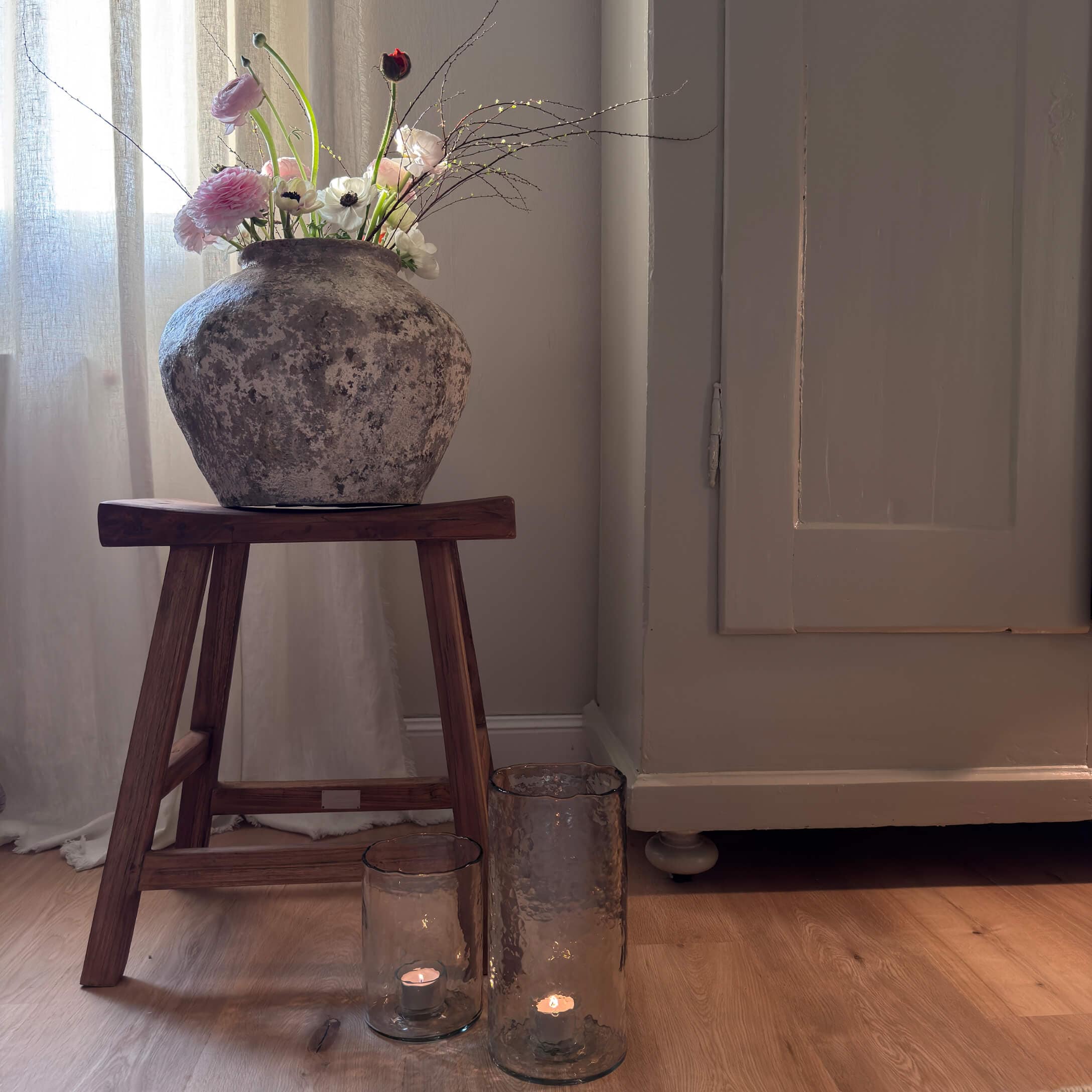 Rustikale Steinvase mit Blumen auf einem Holzschemel neben Kerzen in Glaslaternen