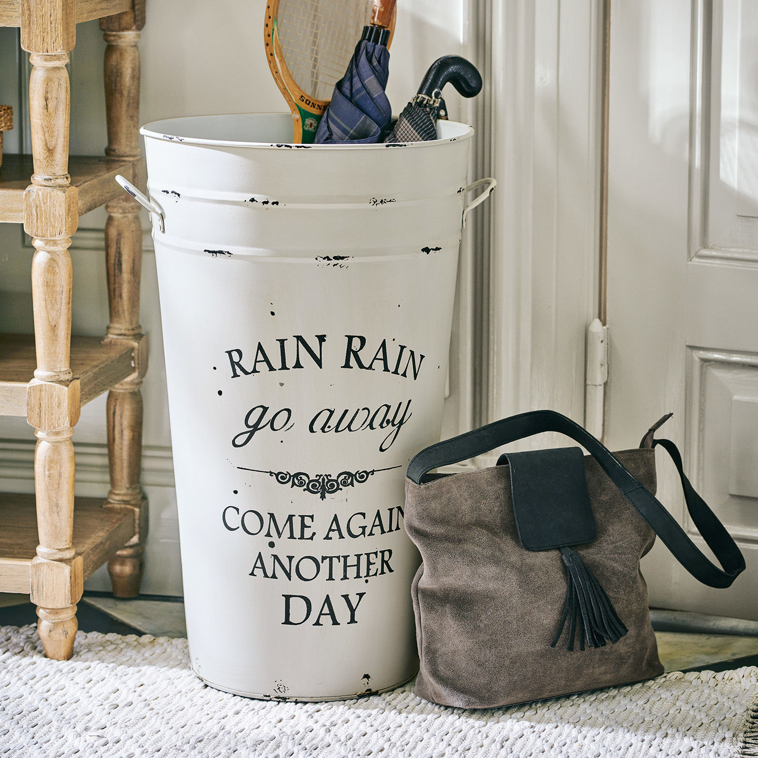 Schirmständer Rain Weisser Metall-Regenschirmständer mit Schriftzug und braune Wildledertasche neben der Türe auf dem Teppich