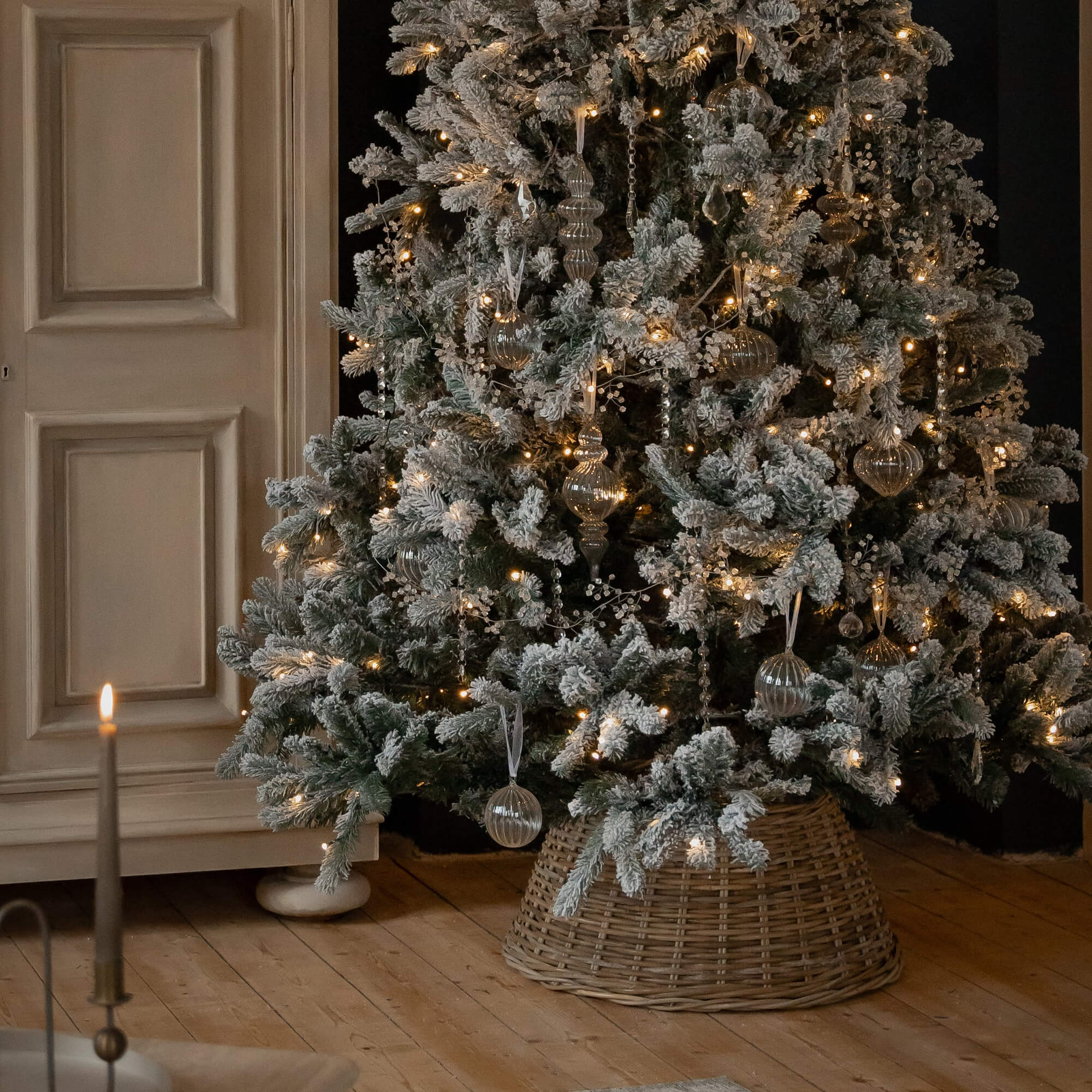 Schneebedeckter Weihnachtsbaum mit Glasornamenten und warmen Lichterketten auf Korbständer