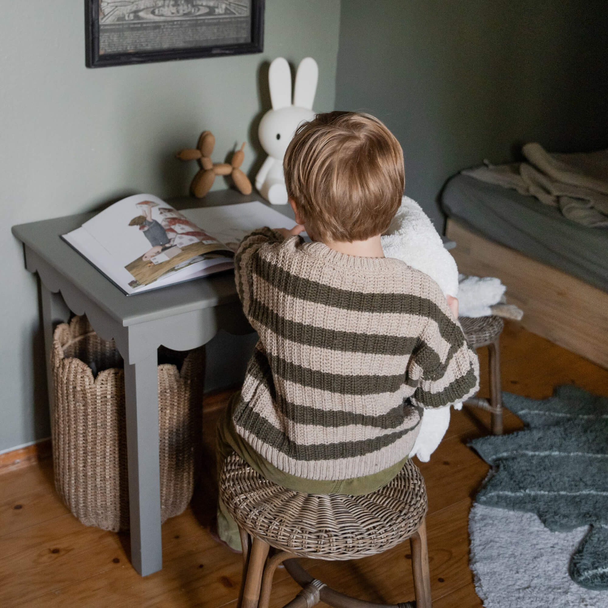 Kind im beige-grünen Pullover sitzt auf Rattanhocker an grauem Holztisch mit Buch und Hasenlampe.