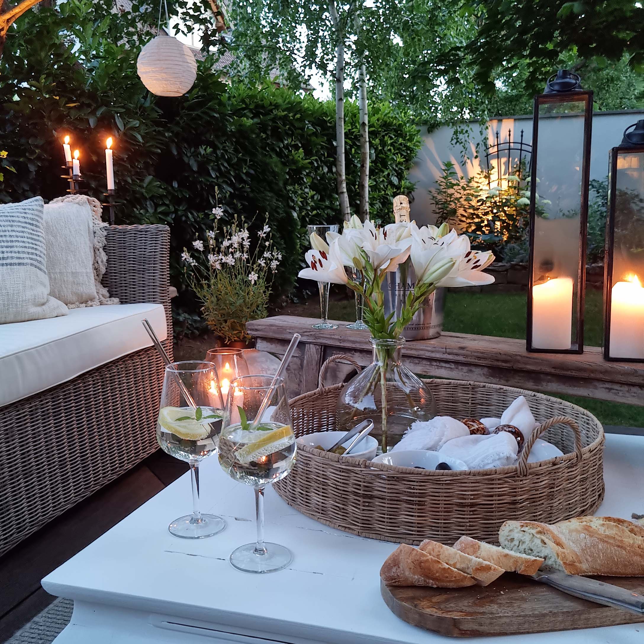 Gemütlicher Garten mit Korbsofa, weissen Lilien und Laternen auf weissem Holztisch