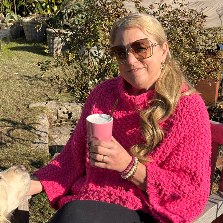 Frau im pinken grob gestrickten Pullover hält einen rosa Becher draussen an einem sonnigen Tag, mit einem Hund neben sich.