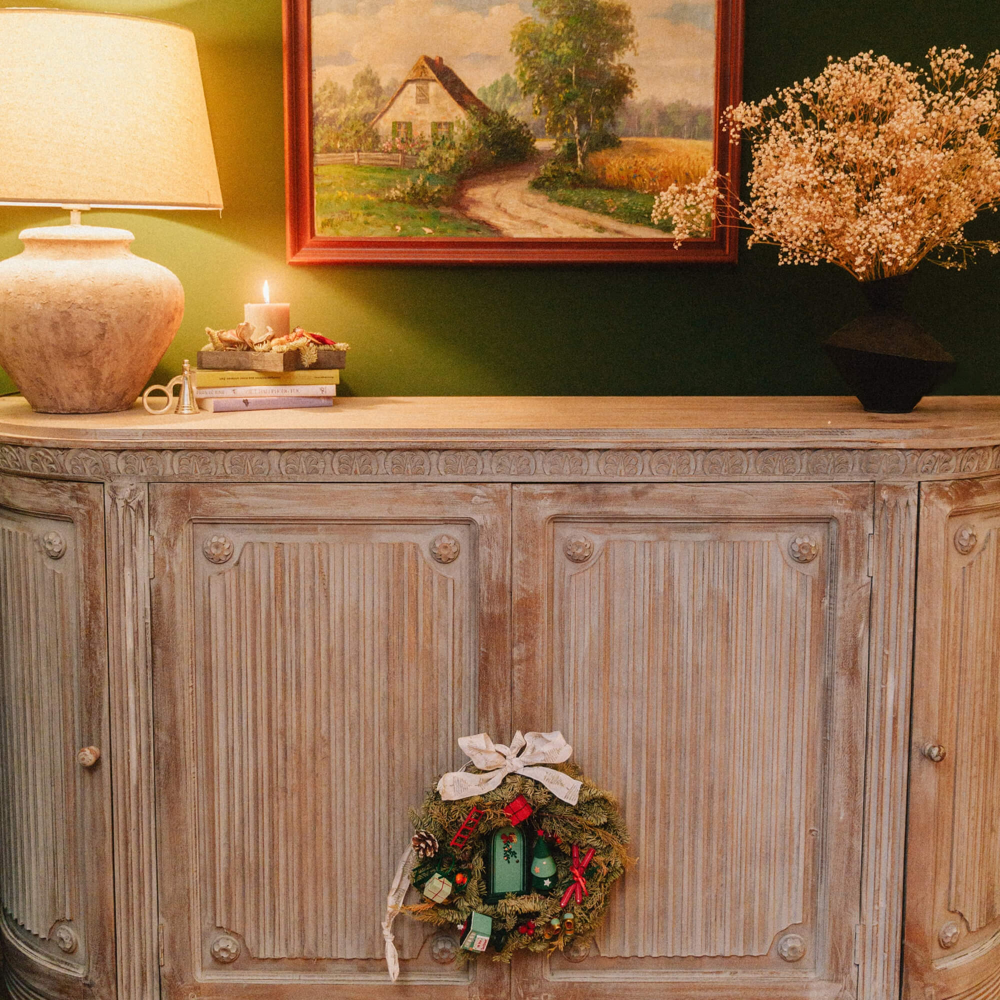 Rustikale Holzkommode mit Weihnachtskranz, Lampe und Landschaftsgemälde an grüner Wand