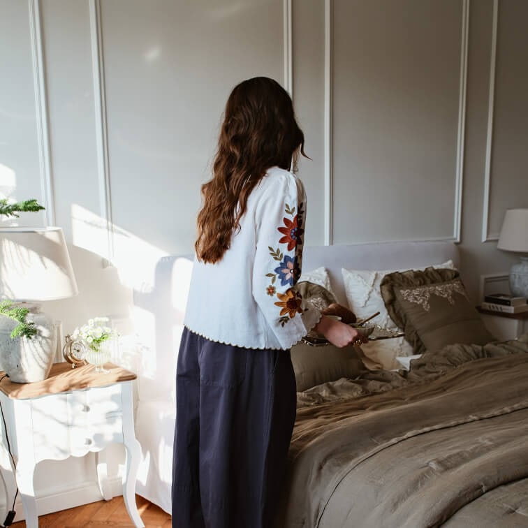 Frau in weisser Bluse mit bunten Stickereien an den Ärmeln im Schlafzimmer mit brauner Bettwäsche und Nachttischlampe