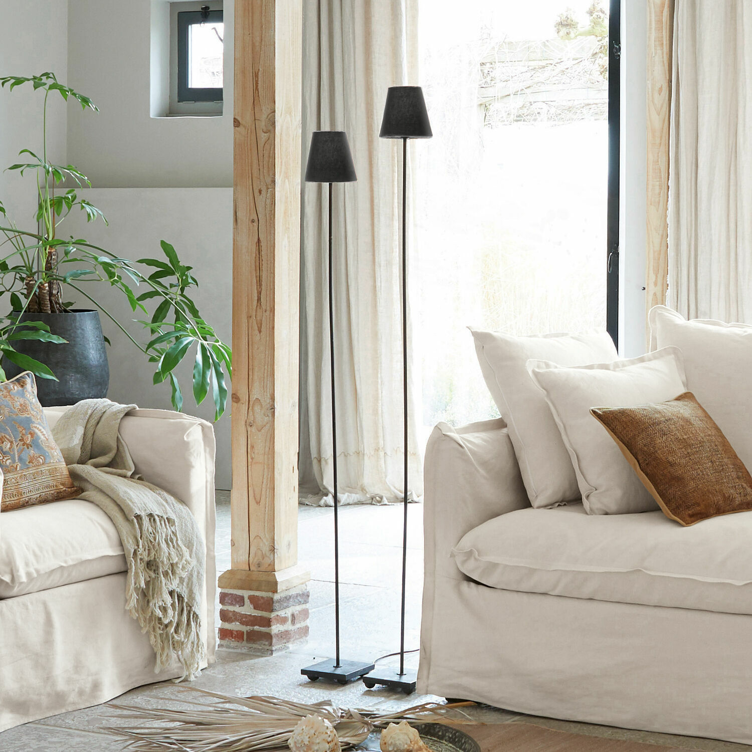 Zwei schwarze Stehlampen aus Metall im hellen Wohnzimmer mit beigem Sofa und grüner Pflanze.