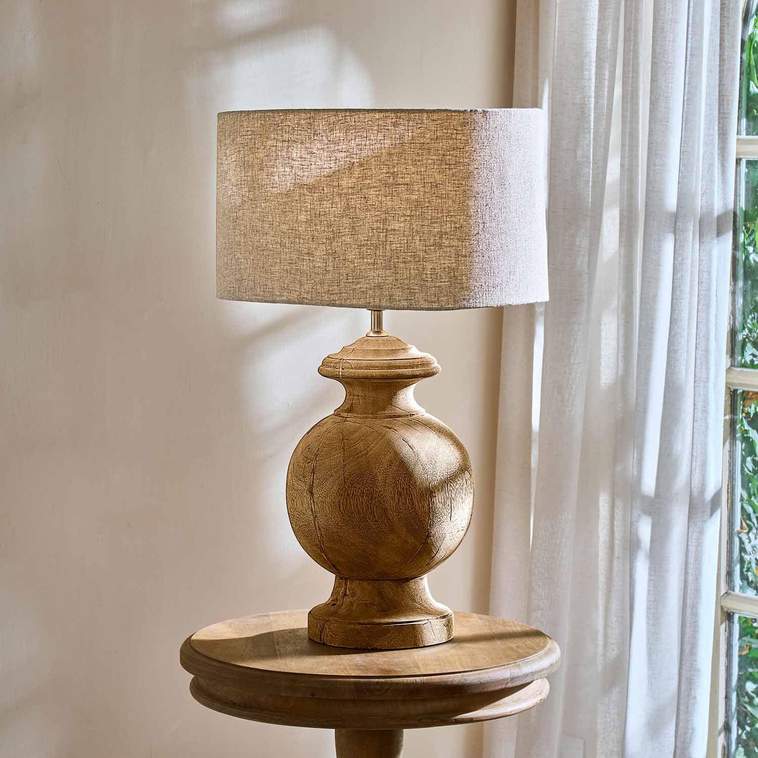 Tischlampe mit runder Holzbasis und beigem Stoffschirm auf rundem Holztisch am Fenster