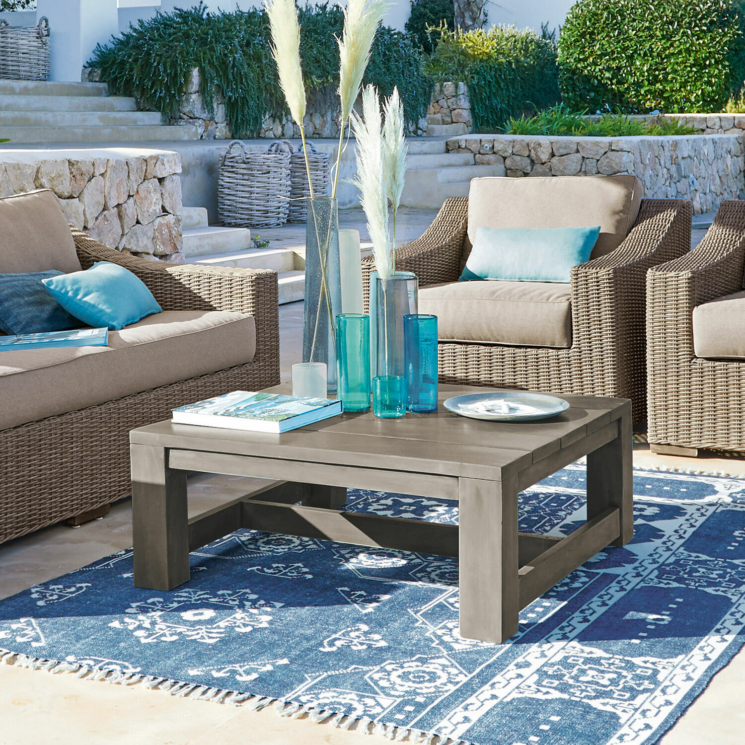 Couchtisch Malden Grauer Couchtisch aus Holz auf blau gemustertem Teppich, umgeben von Korbsesseln mit blauen Kissen.