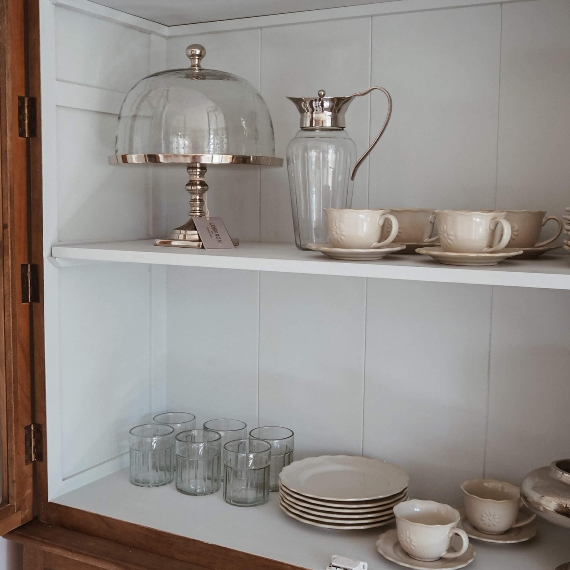 Set aus cremefarbenen Keramiktassen und Tellern mit Glas-Krug und Kuchenhaube im weissen Schrank