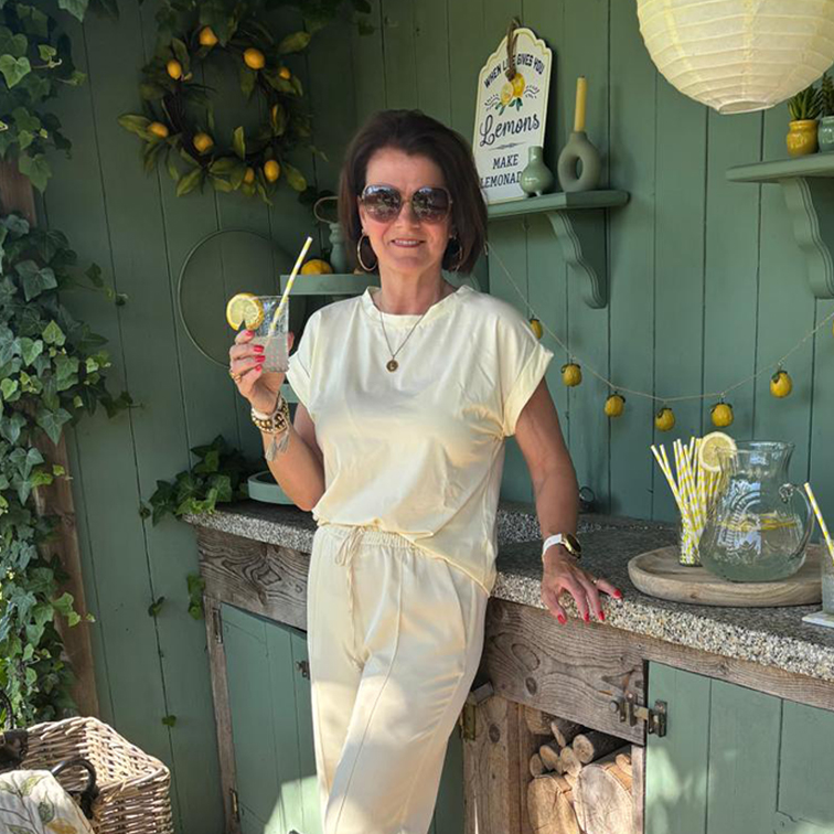 Frau im cremefarbenen Outfit entspannt mit Limonade vor grünem Wandhintergrund mit Zitronendekor