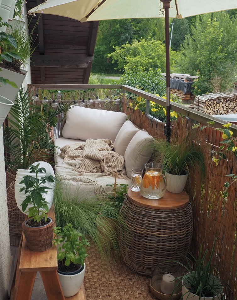 Gemütlicher Balkon mit beigem Sofa, Kissen, Rattantisch, Pflanzen und Sonnenschirm zur Entspannung