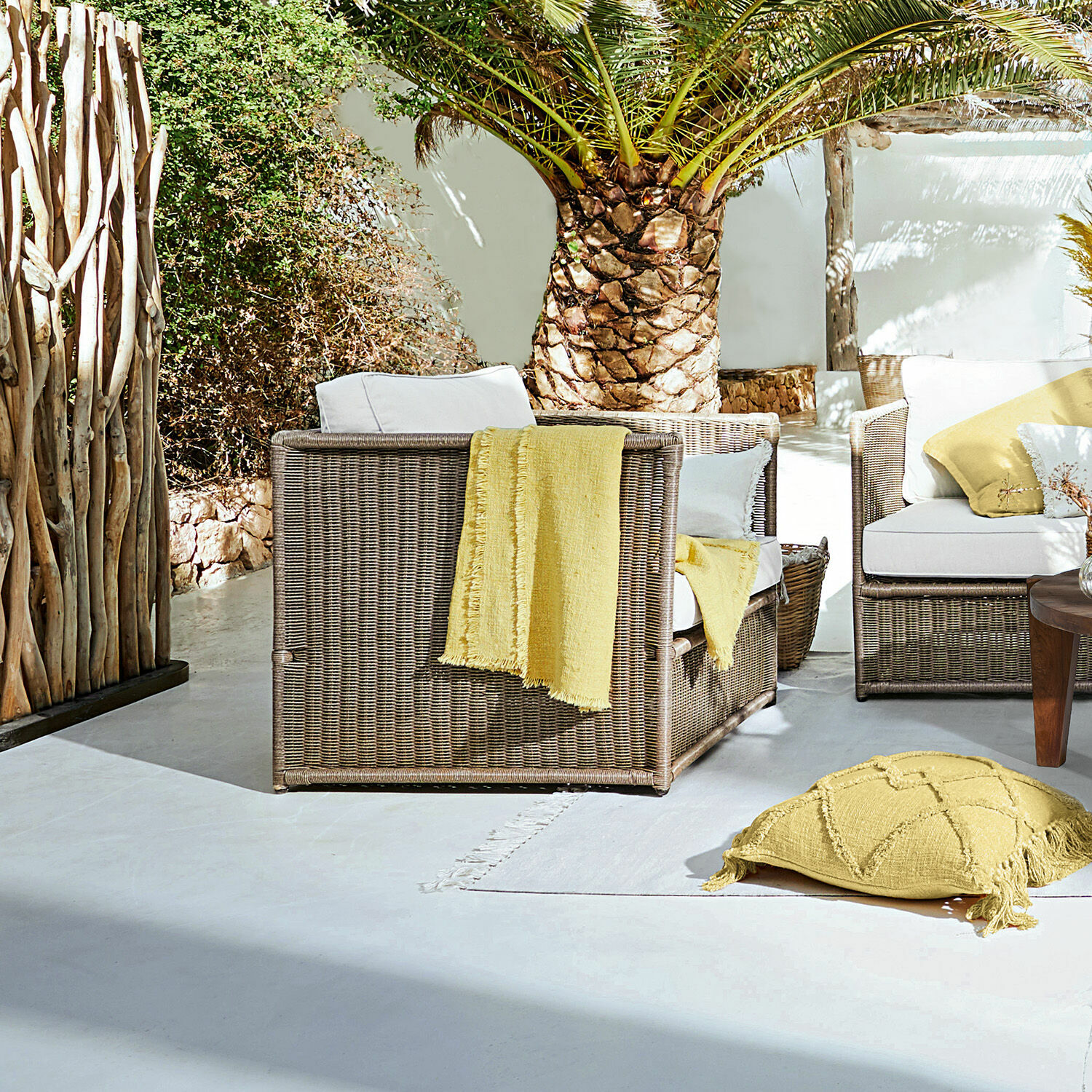 Sessel Lesmont Braunes Rattan-Gartenmöbel-Set mit weissen Kissen und gelben Akzenten auf sonniger Terrasse