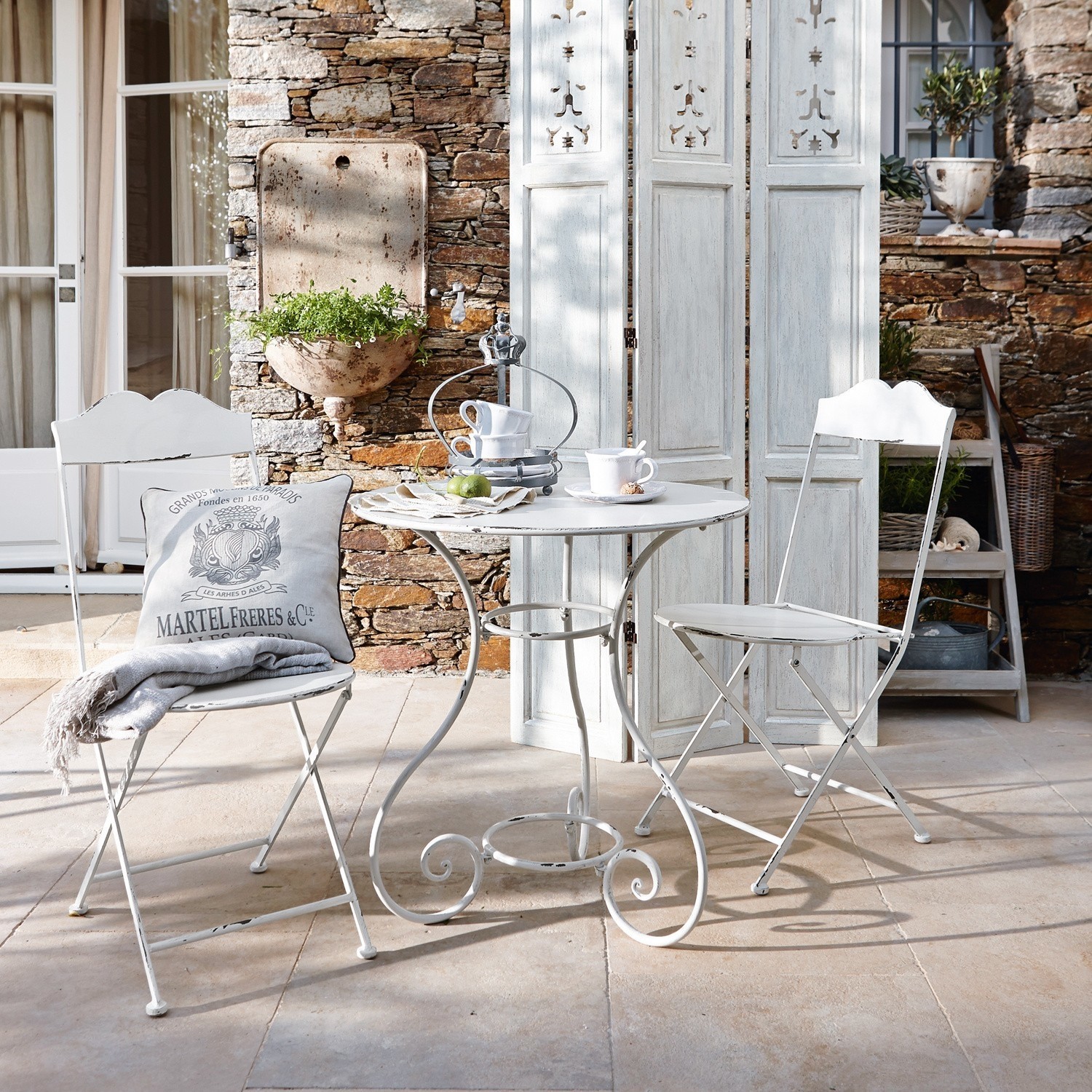 Sitzgruppe Iteuil Weisser Metall-Bistrotisch mit zwei Klappstühlen im Vintage-Stil auf Terrasse mit Steinmauer