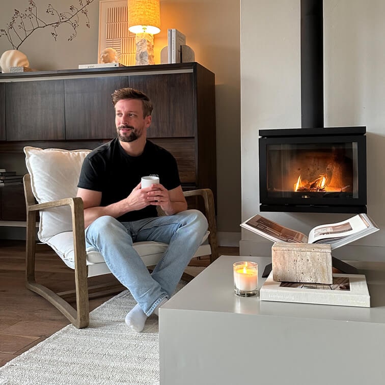 Mann sitzt auf Holzstuhl mit weissem Kissen, hält Tasse, Kamin und Kerze auf dem Tisch im Wohnzimmer