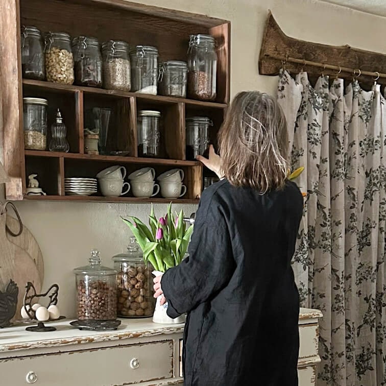 Frau in schwarzem Kleid greift nach Glas auf Holzregal in rustikaler Küche mit Tulpenstrauß
