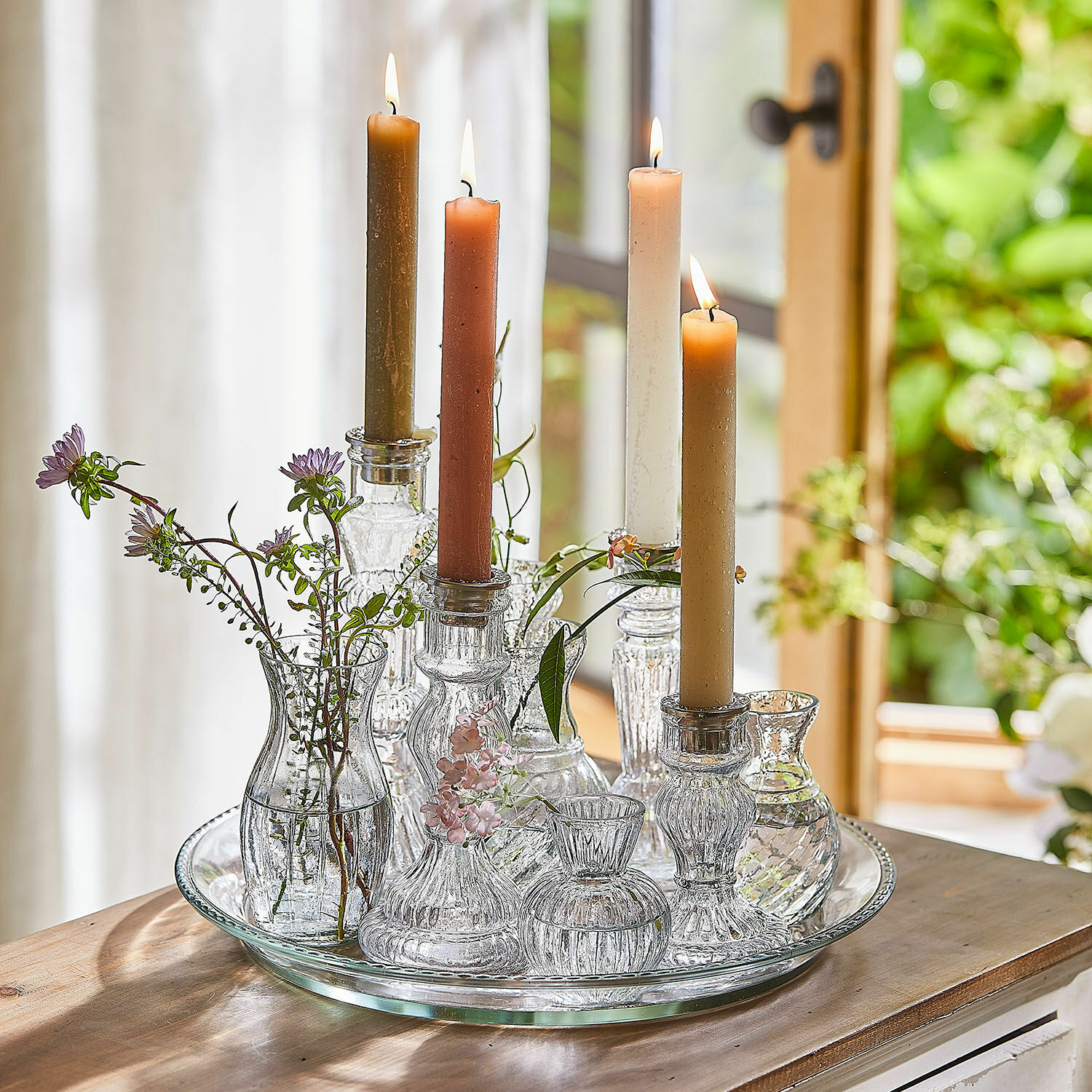 Tablett mit Kerzenständer und Vase Hilevor Gläserne Kerzenhalter und Väschen mit farbigen Kerzen und Blumen auf einem Glastablett auf dem Holztisch