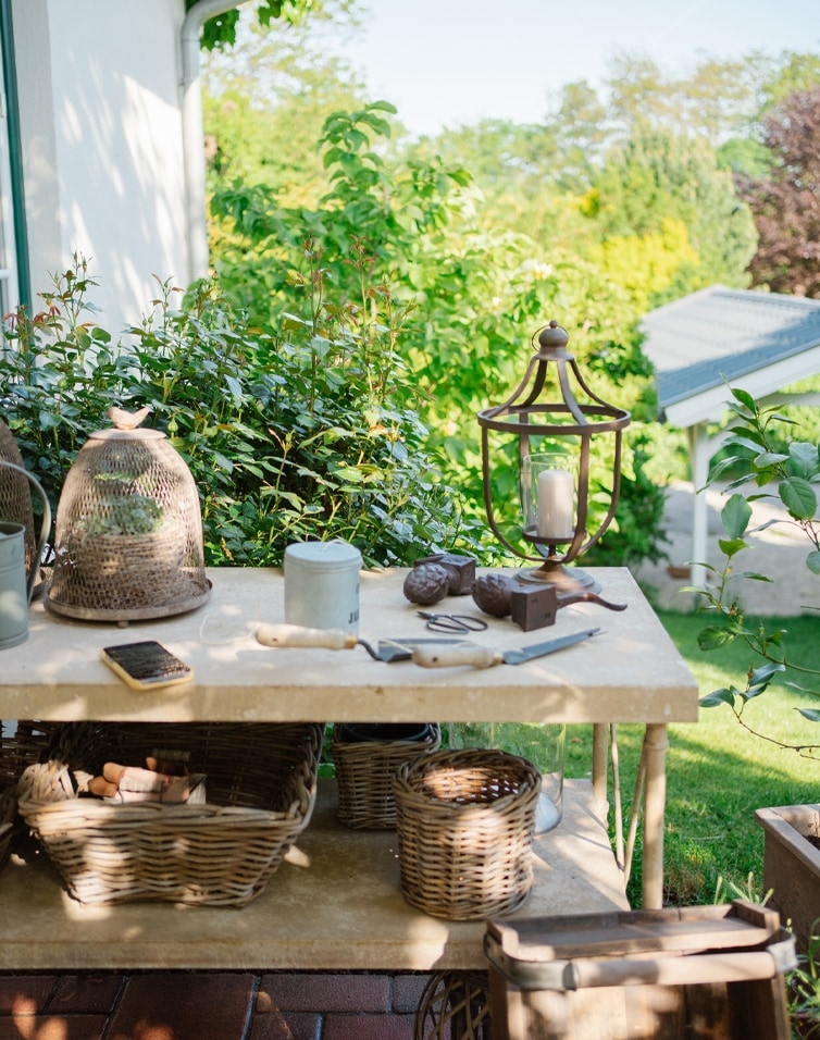 Gartentisch aus Stein mit Weidenkörben, Gartenwerkzeugen und Kerzenlaterne im Grünen.