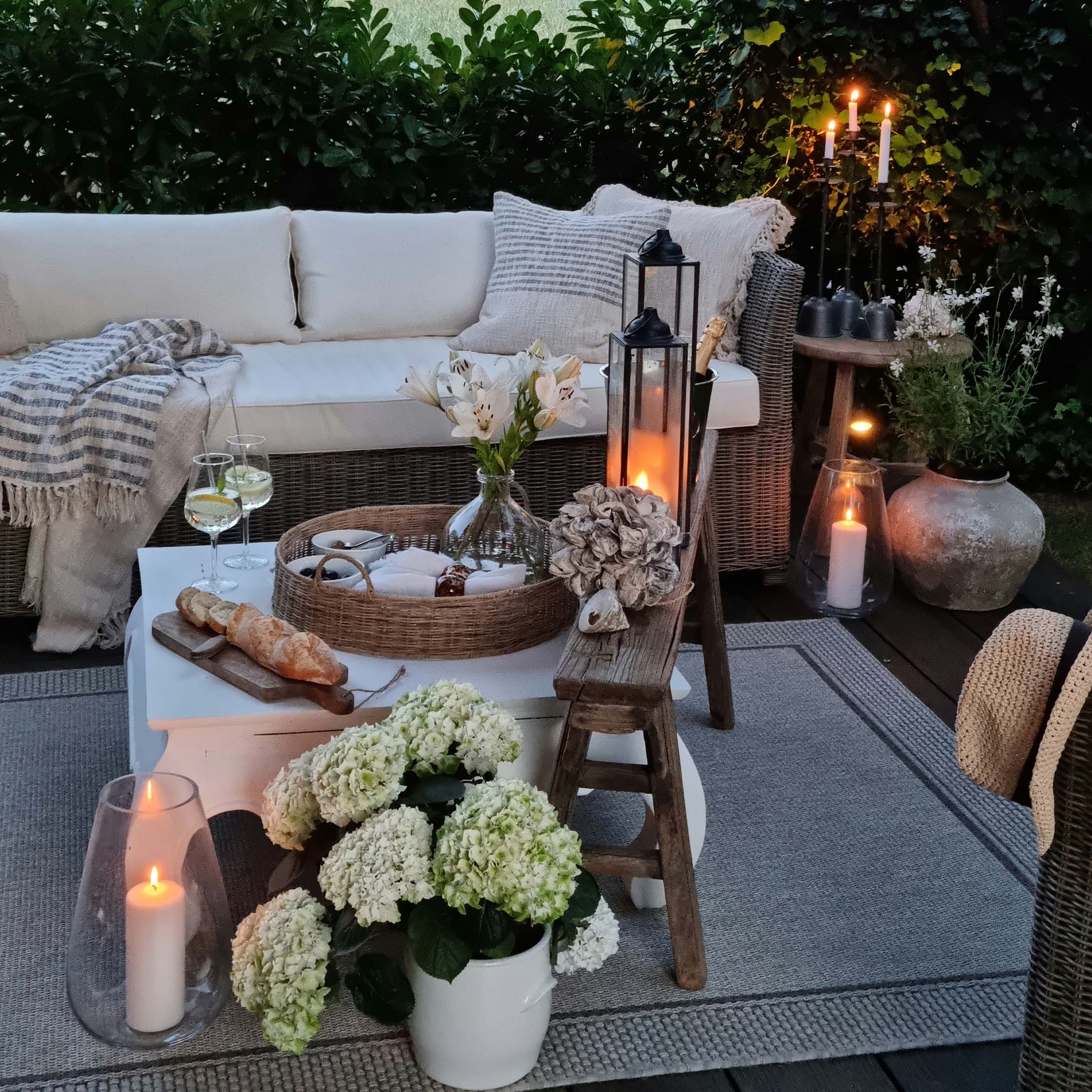 Gemütliche Gartenecke mit Rattansofa, weissen Kissen und Kerzen auf Holzterrasse