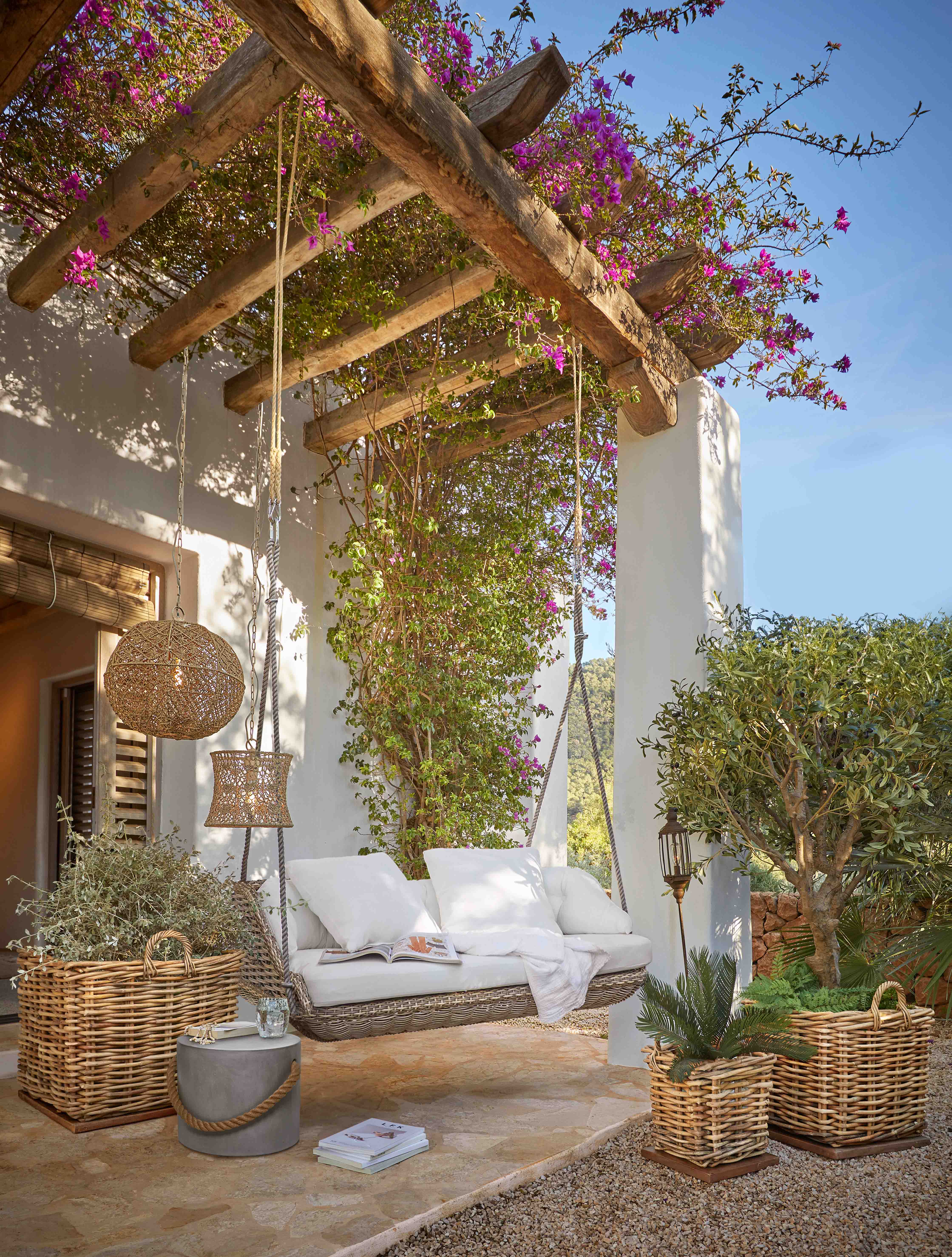 Wiklingschaukel mit weissen Kissen unter einer Holzpergola mit blühenden pinken Blumen