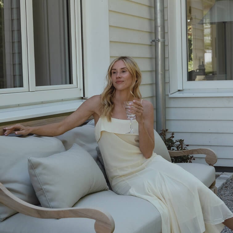 Frau im cremefarbenen Kleid sitzt entspannt auf hellgrauem Sofa im Freien und hält ein Weinglas