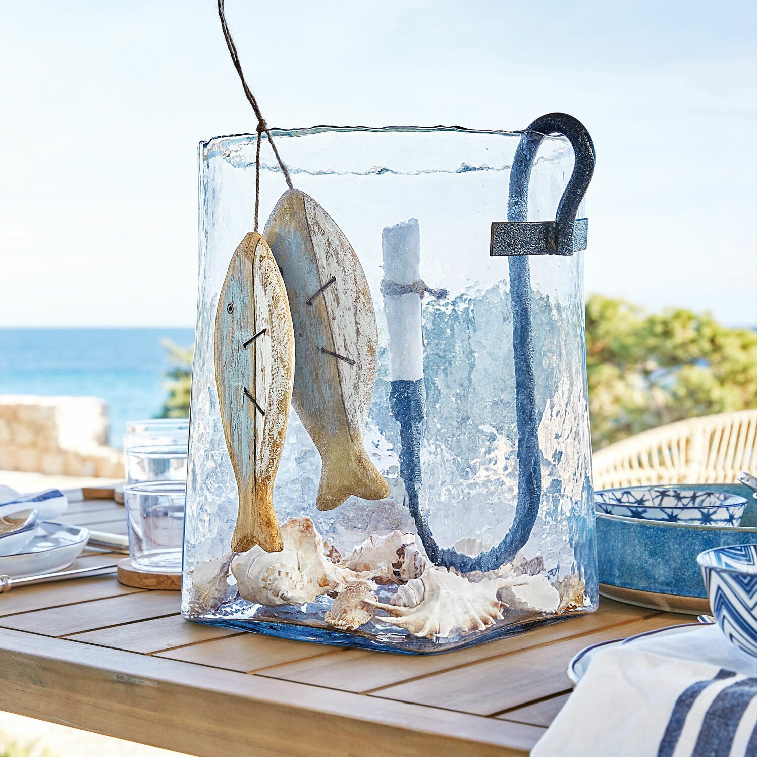 Glas-Lampion mit Metallgriff, Holzfisch-Dekor und Muscheln auf Holztisch auf Terrasse mit Meerblick