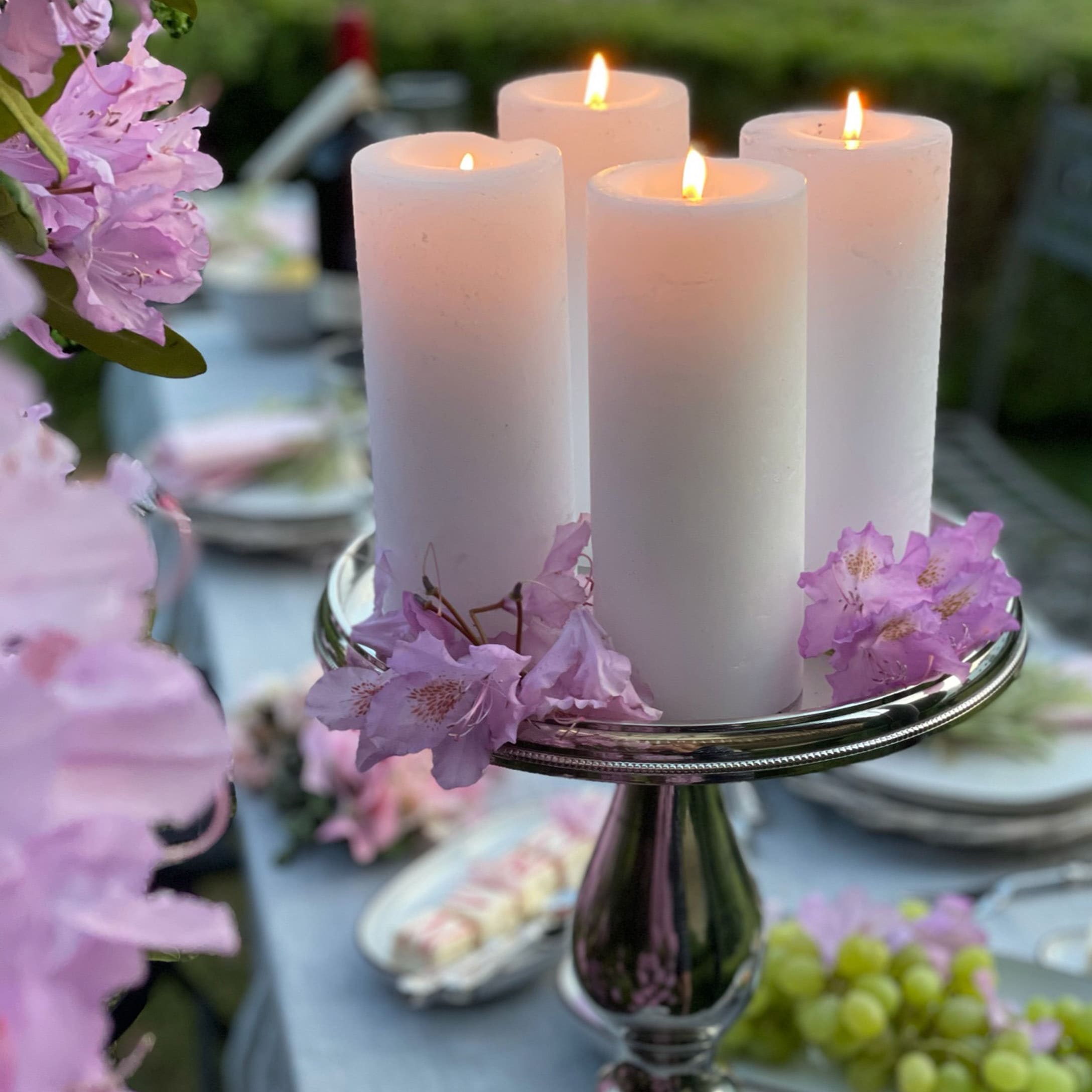 Vier weisse Kerzen auf silbernem Kerzenhalter mit rosa Blüten auf Gartentisch