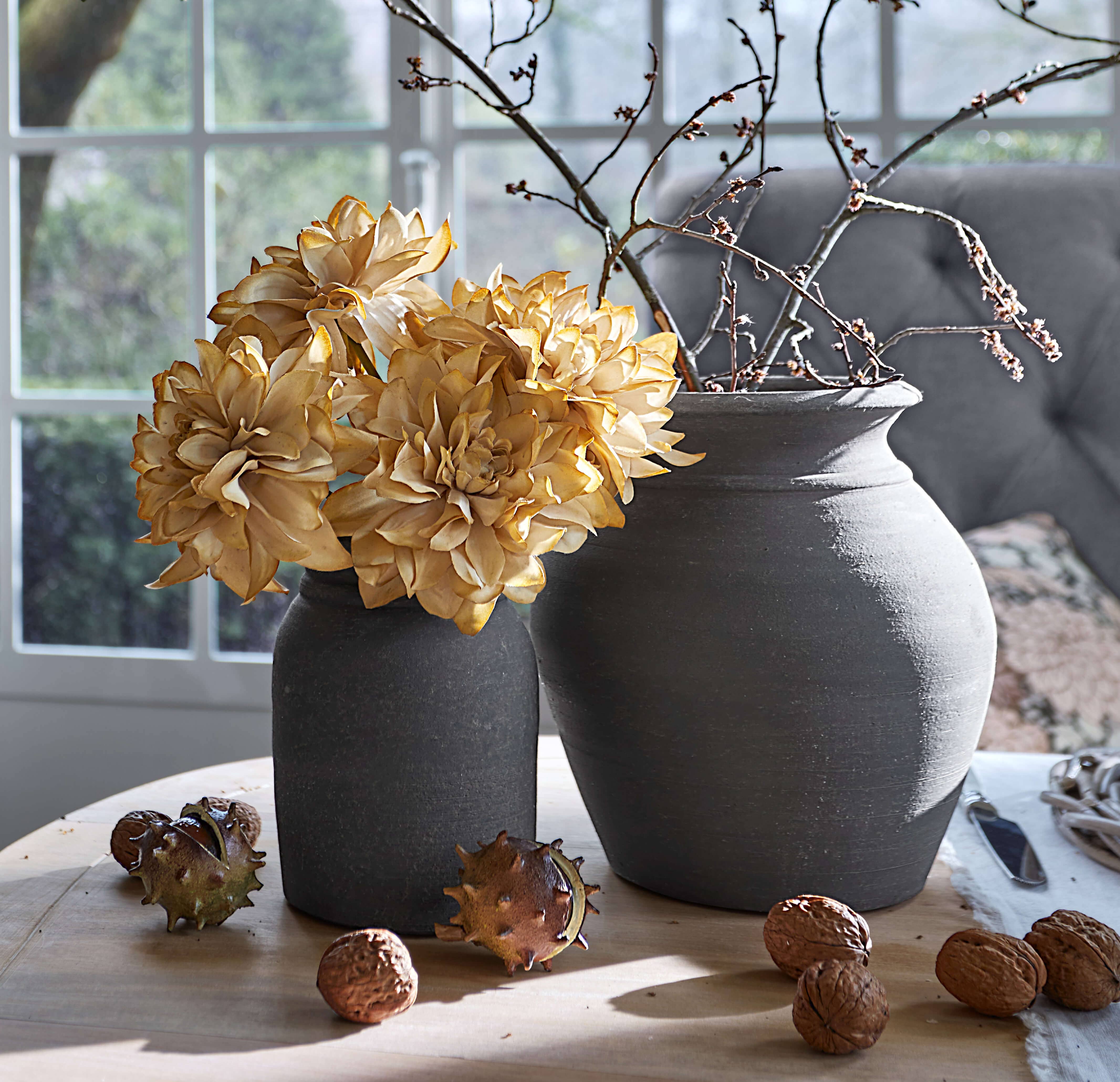 Zwei graue Keramikvasen mit getrockneten Blumen und Zweigen auf Holztisch mit Walnüssen