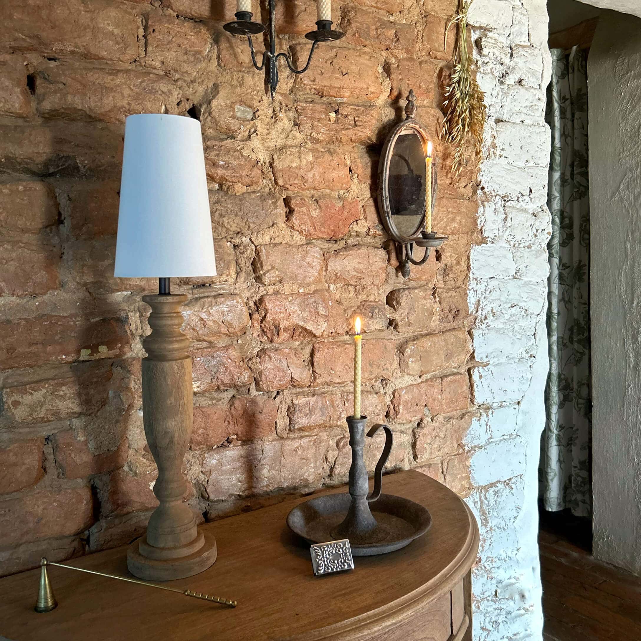 Holztisch mit Holzlampe mit weissem Schirm und Metallkerzenhalter mit brennender Kerze vor Ziegelwand