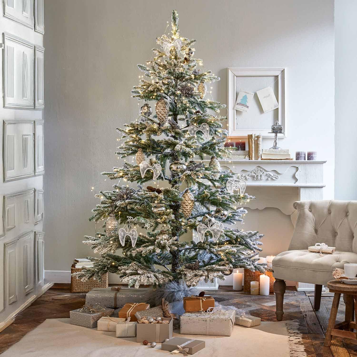 Schneebedeckter Weihnachtsbaum mit weissen Engelsanhängern und goldenen Zapfen in gemütlichem Wohnzimmer mit Kamin
