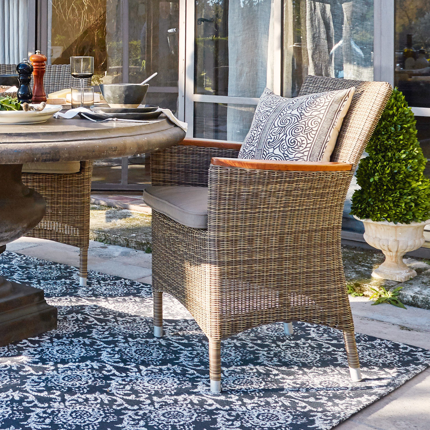 Sessel Casper Rattanstuhl in Naturfarbe mit Polster und Holzarmlehnen auf der Terrasse neben dem Tisch